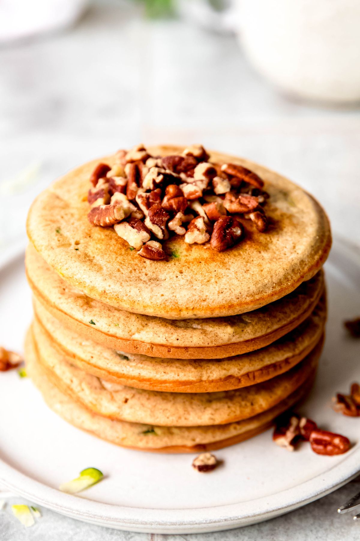 45 degree angle closeup shot of a round white plate with a stack of fluffy zucchini pancakes topped with chopped pecans.