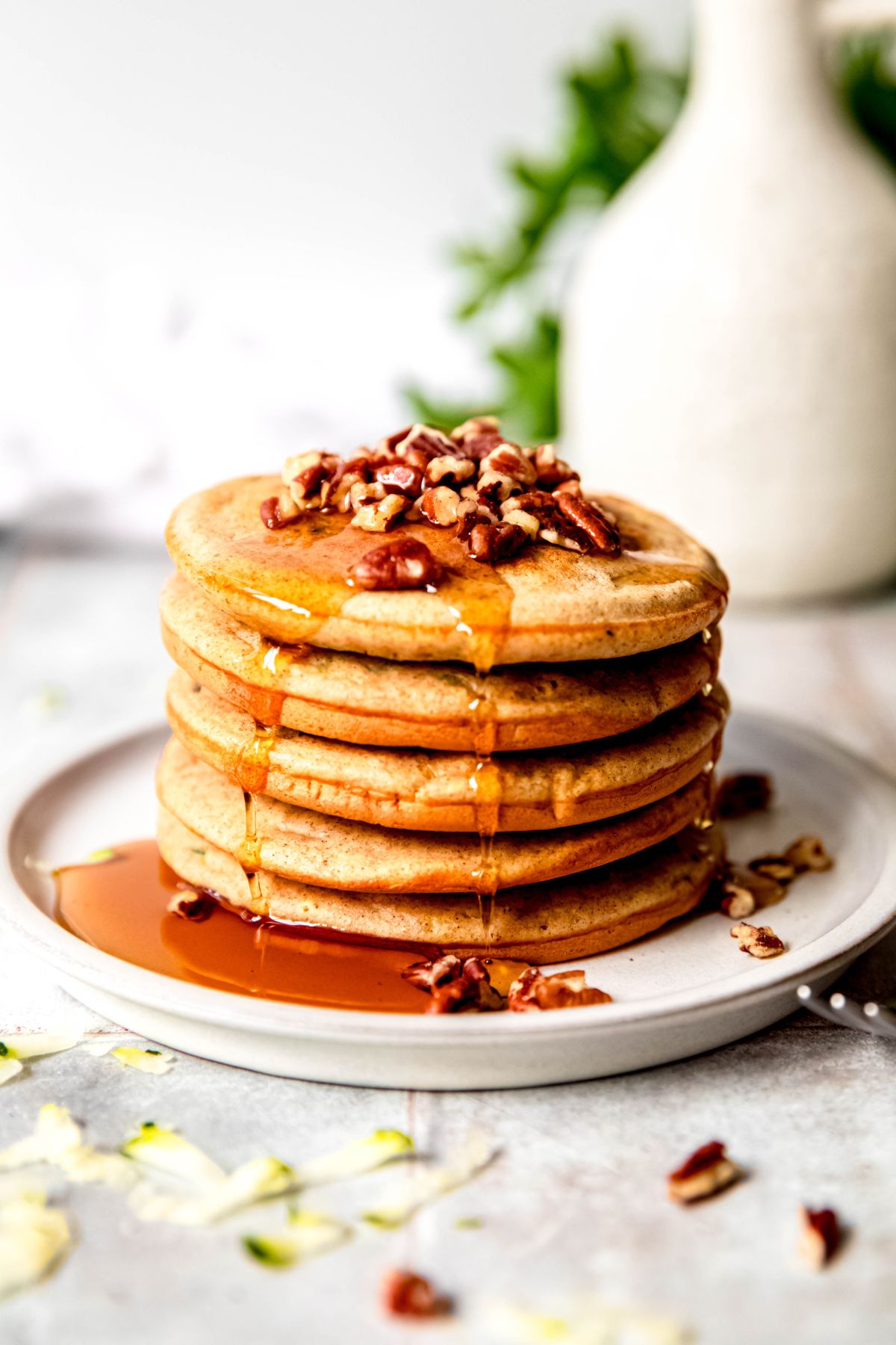 stack of 5 fluffy protein zucchini pancakes on a white plate topped with chopped pecans and drizzled with syrup that is dripping down the sides onto the plate.