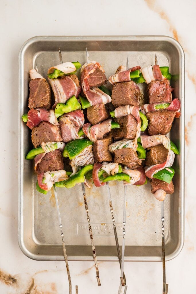 raw marinated beef and bell pepper threaded onto skewers with bacon slices woven through.