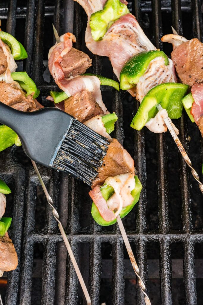 action shot of a silicone pastry brush basting the bacon beef skewers on the grill.