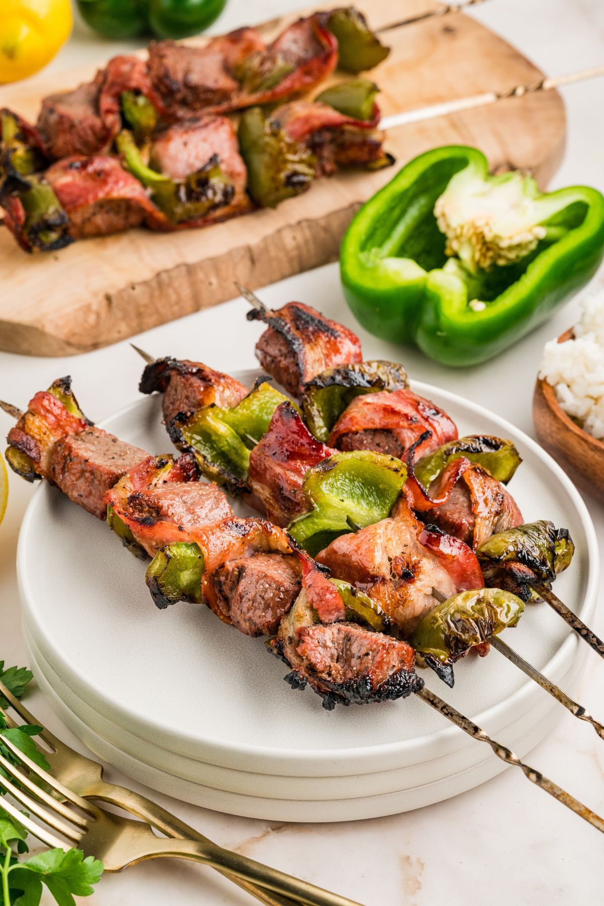 three beef and bacon skewers on the top of a stack of 3 white appetizer plates on a white table with a halved bell pepper, a wooden bowl with rice, and silver forks.