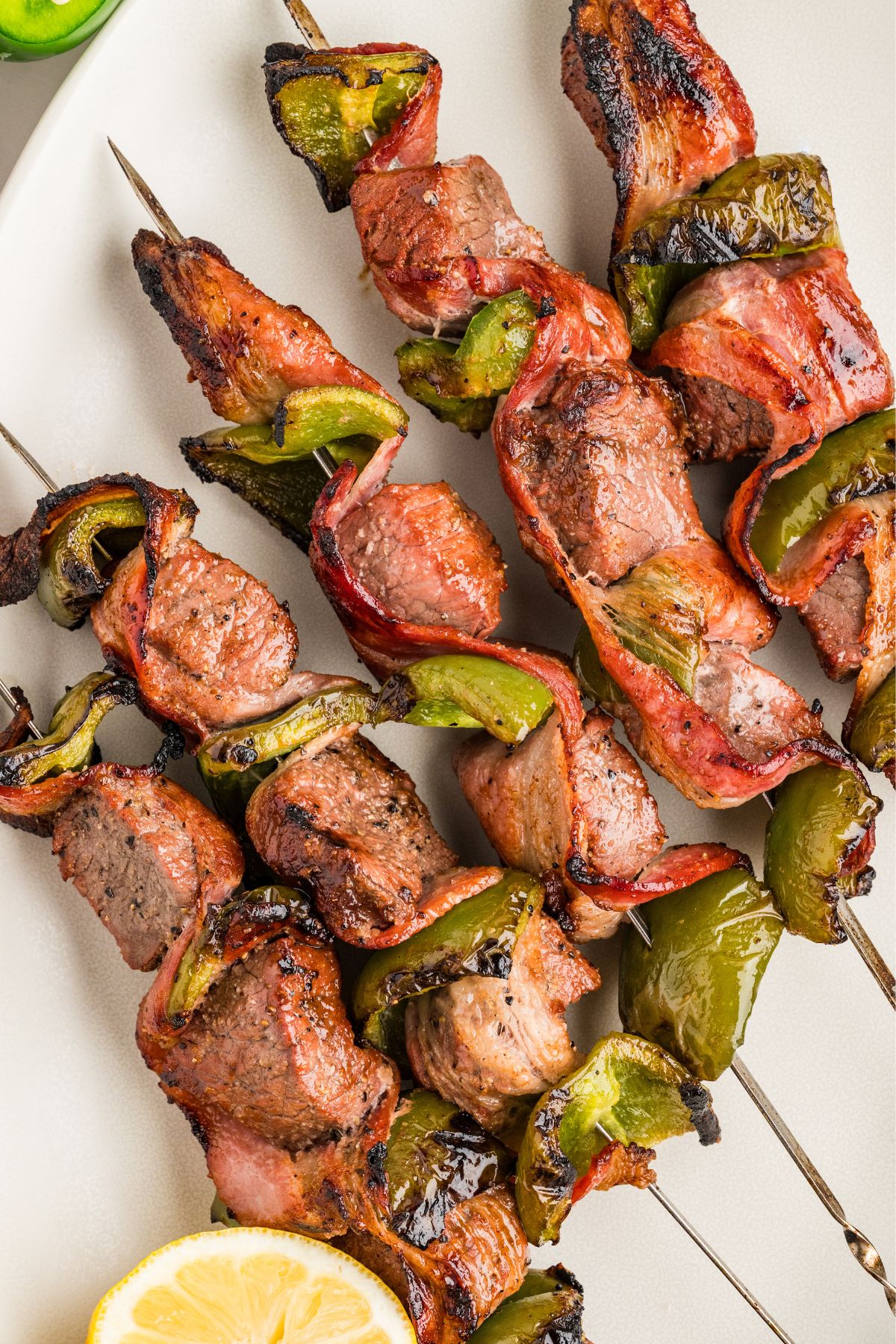 a line of bacon, bell pepper, and beef skewers on a white plate after grilling.