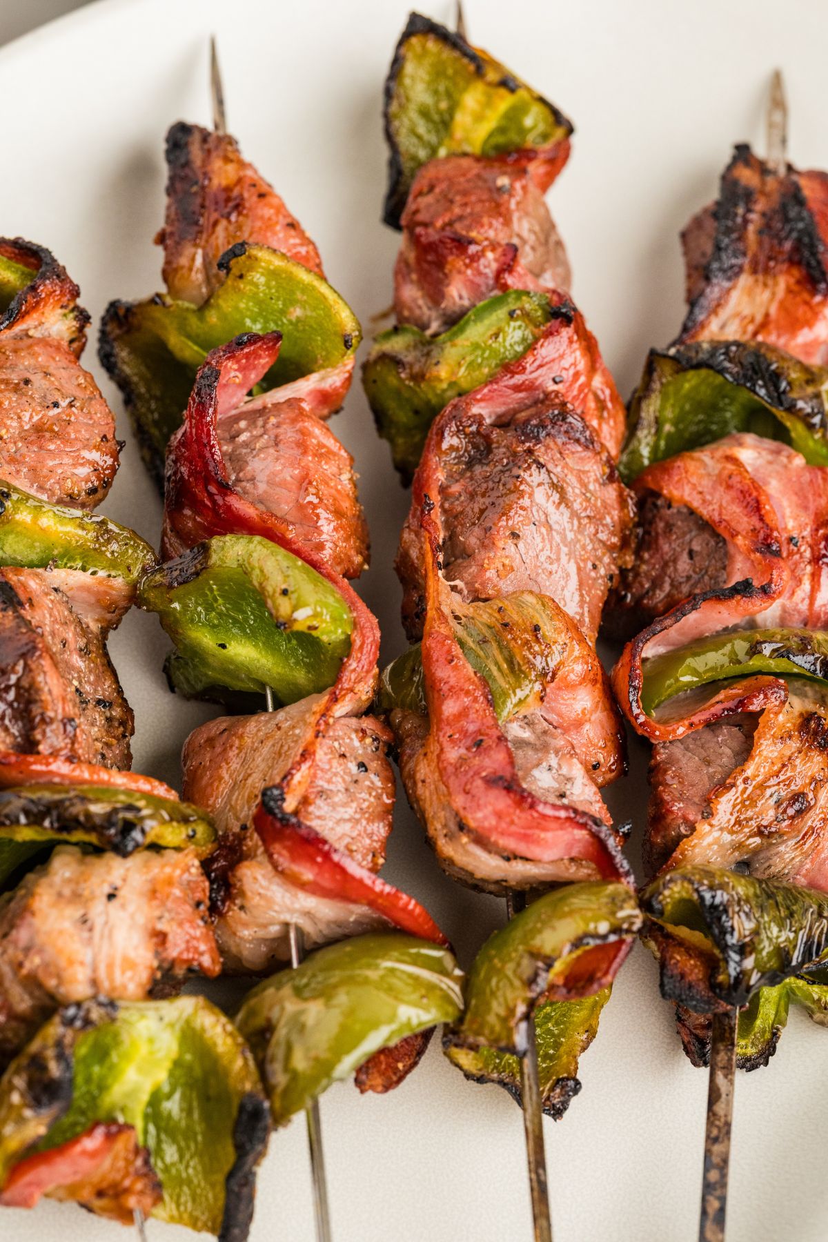 closeup shot of bell pepper, bacon and beef steak skewers on a white plate.