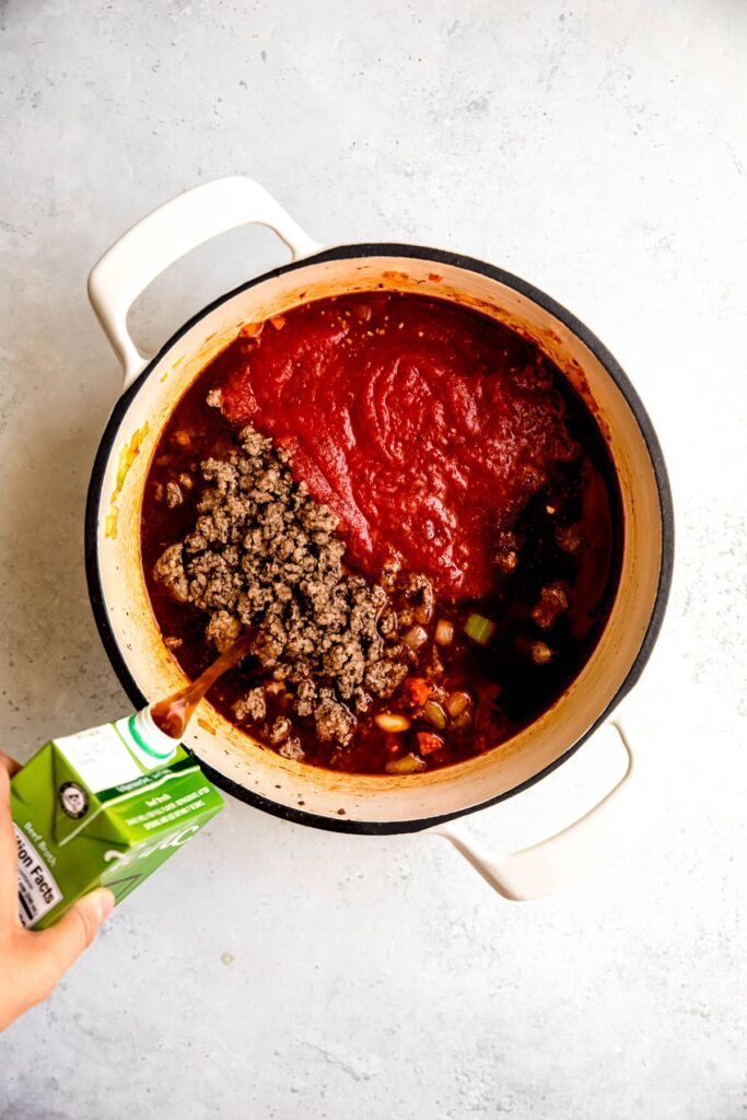 Adding beef broth to a white Dutch oven of ground beef, marinara sauce, and mirepoix.