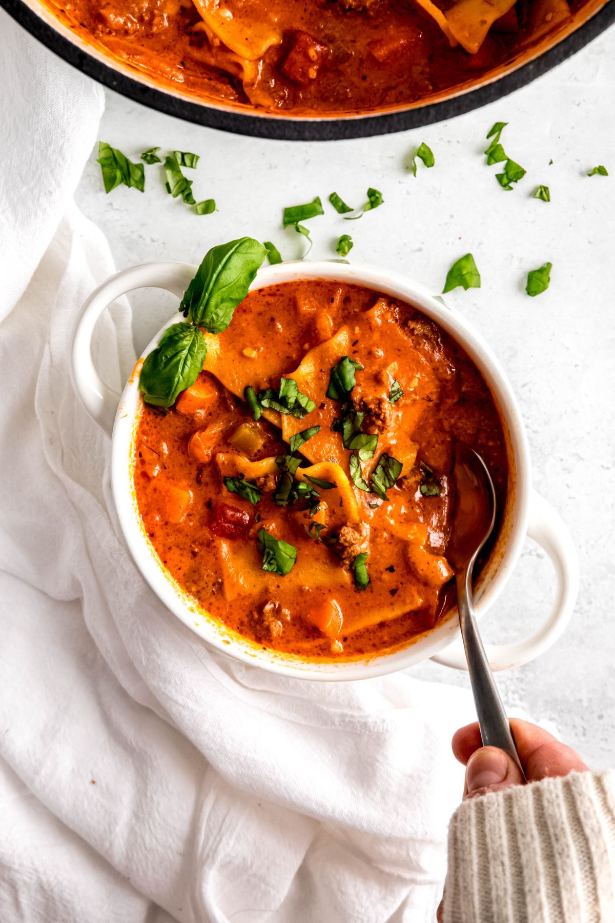An overhead shot of a hand in a sweater taking a scoop of the lasagna soup.