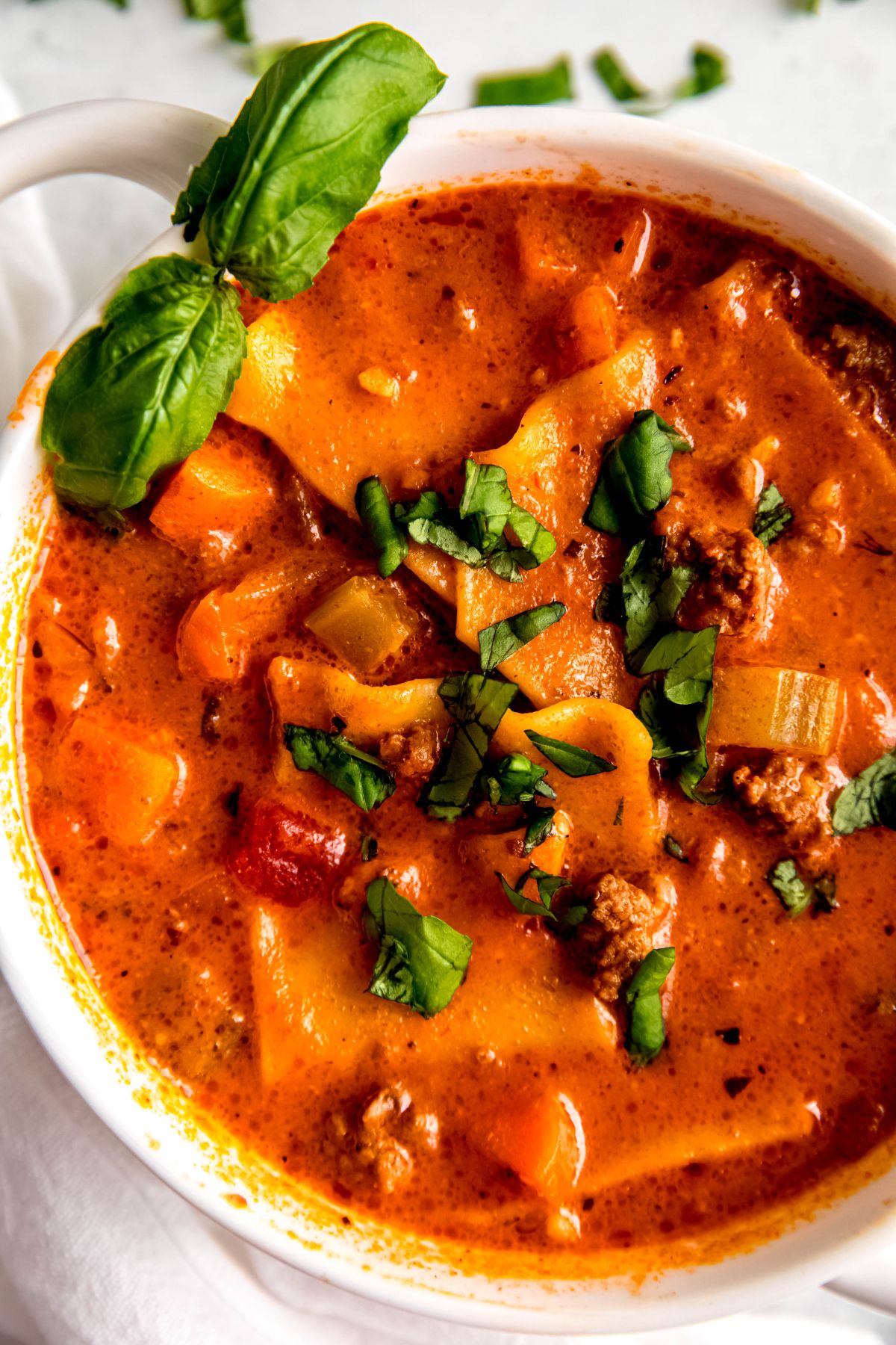 A close-up overhead shot of a bowl of lasagna soup topped with chopped basil.