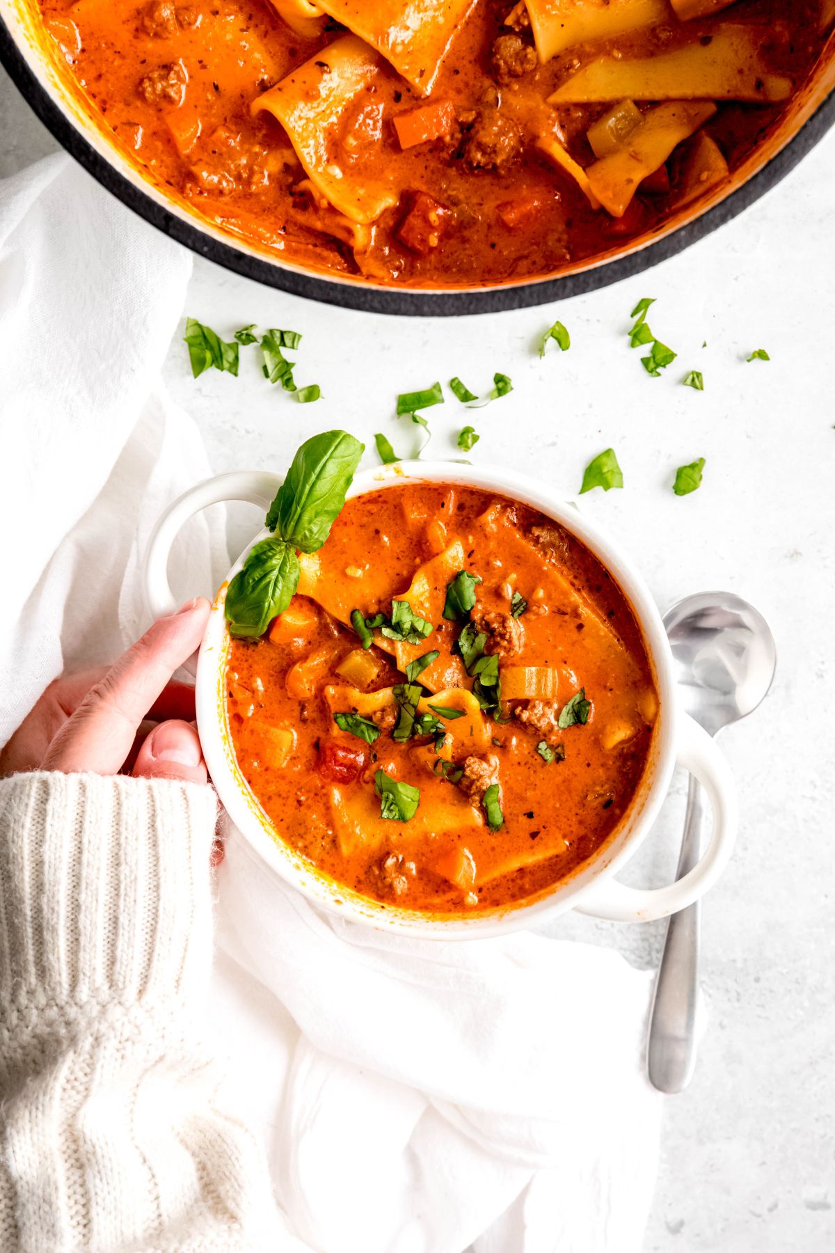 An overhead shot of a hand in a white sweater reaching for a bowl of the lasagna soup.