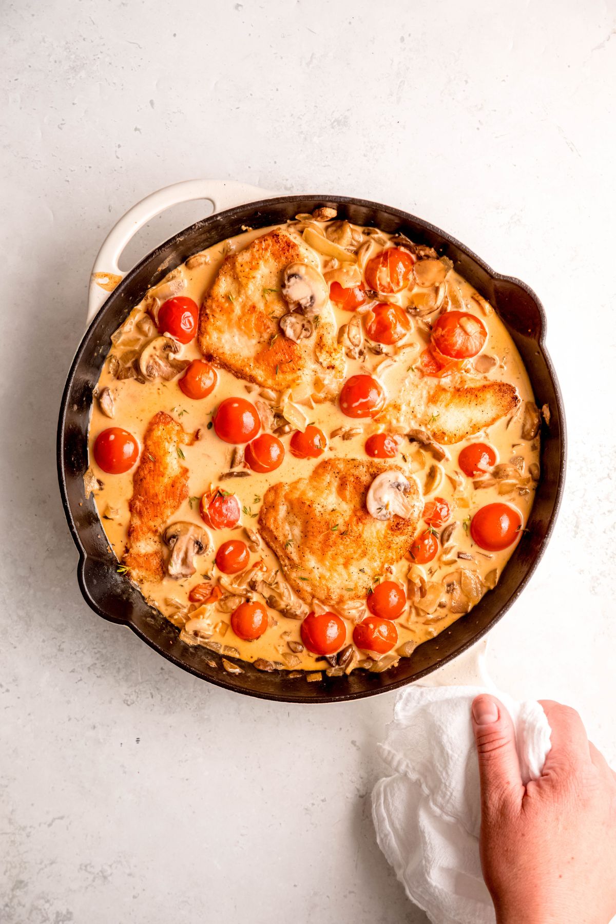 An overhead shot of a hand holding a pan of creamy mushroom chicken with tomatoes.