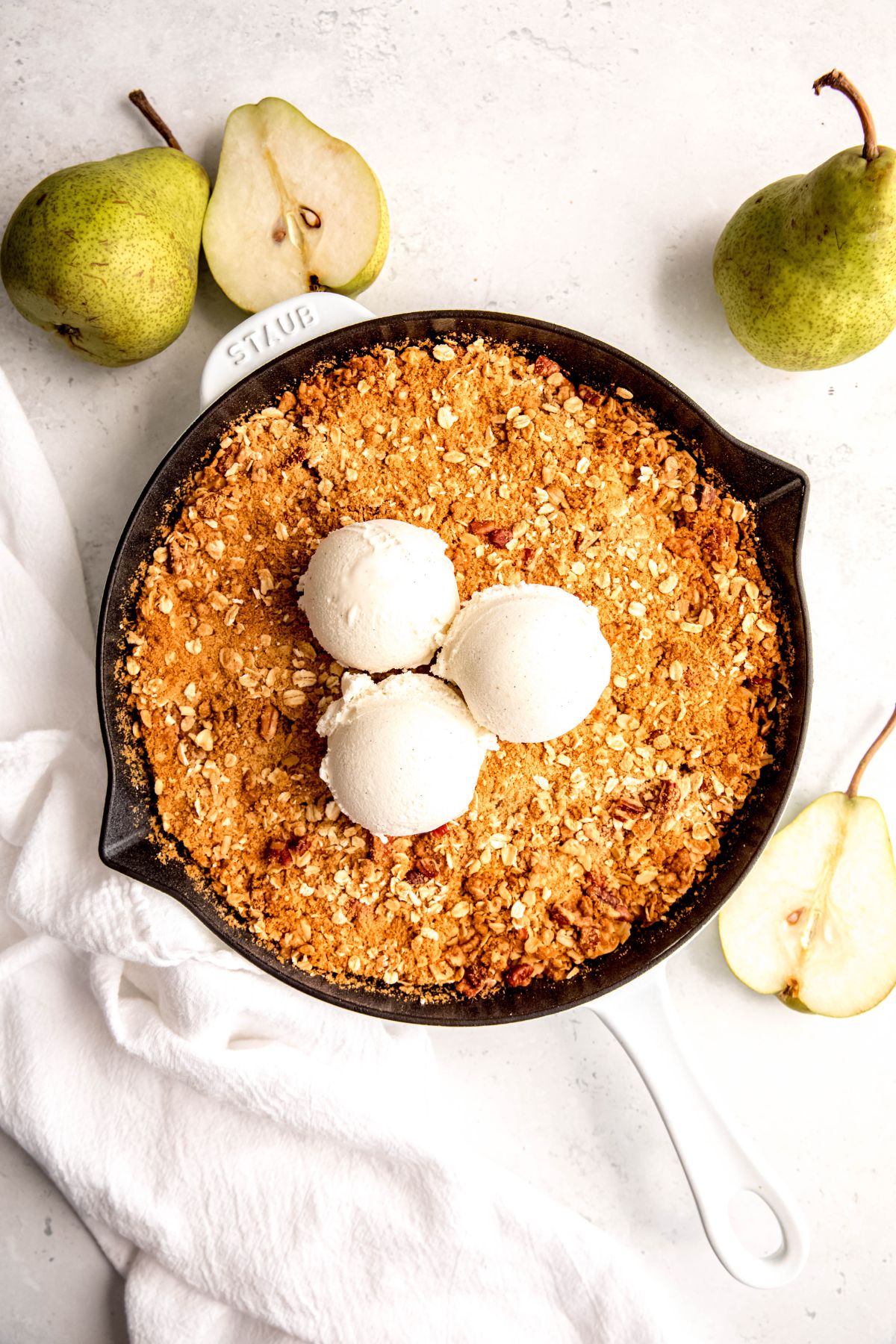 white cast iron skillet filled with healthy pear crisp topped with 3 scoops of vanilla ice cream on a white table with fresh pears.