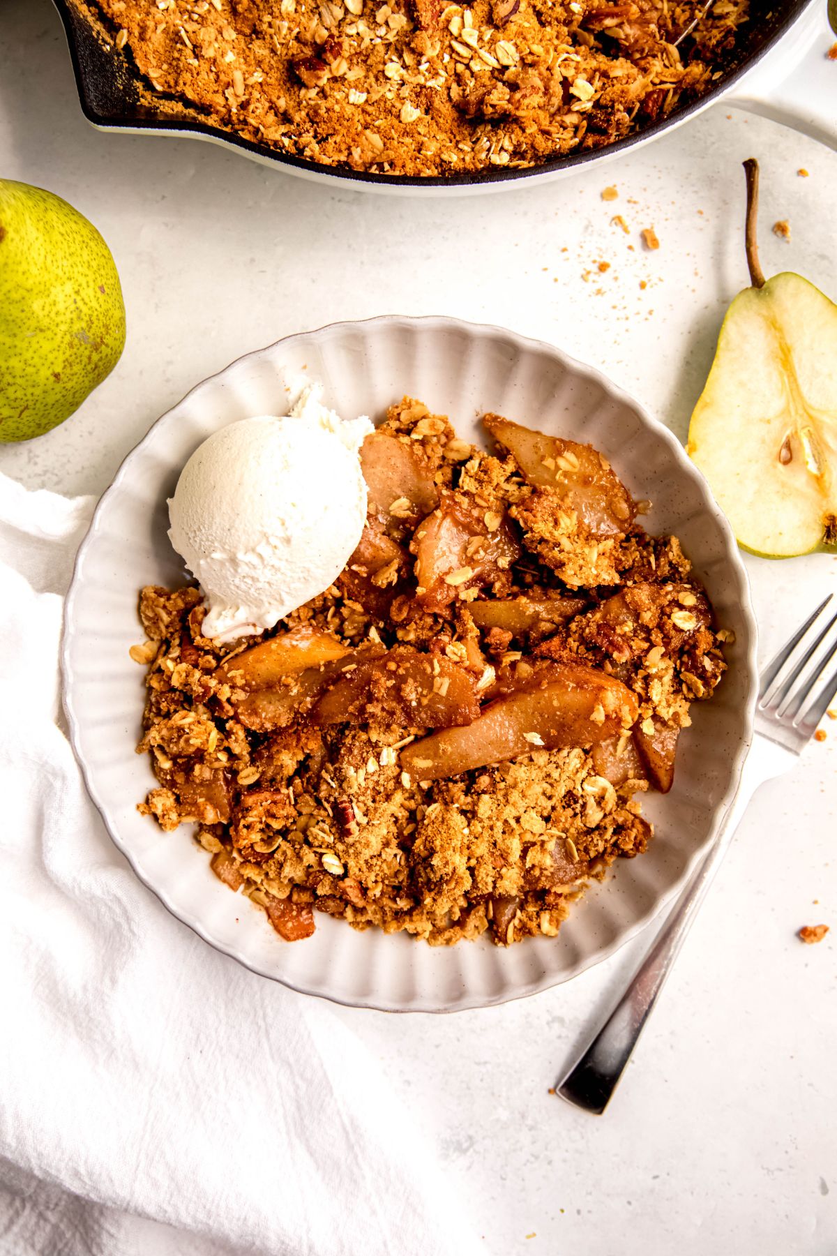 flat lay shot of a white bowl filled with a serving of healthy apple crisp with fresh pears topped with a scoop of vanilla ice cream on a white table with a silver fork and a halved fresh pear.
