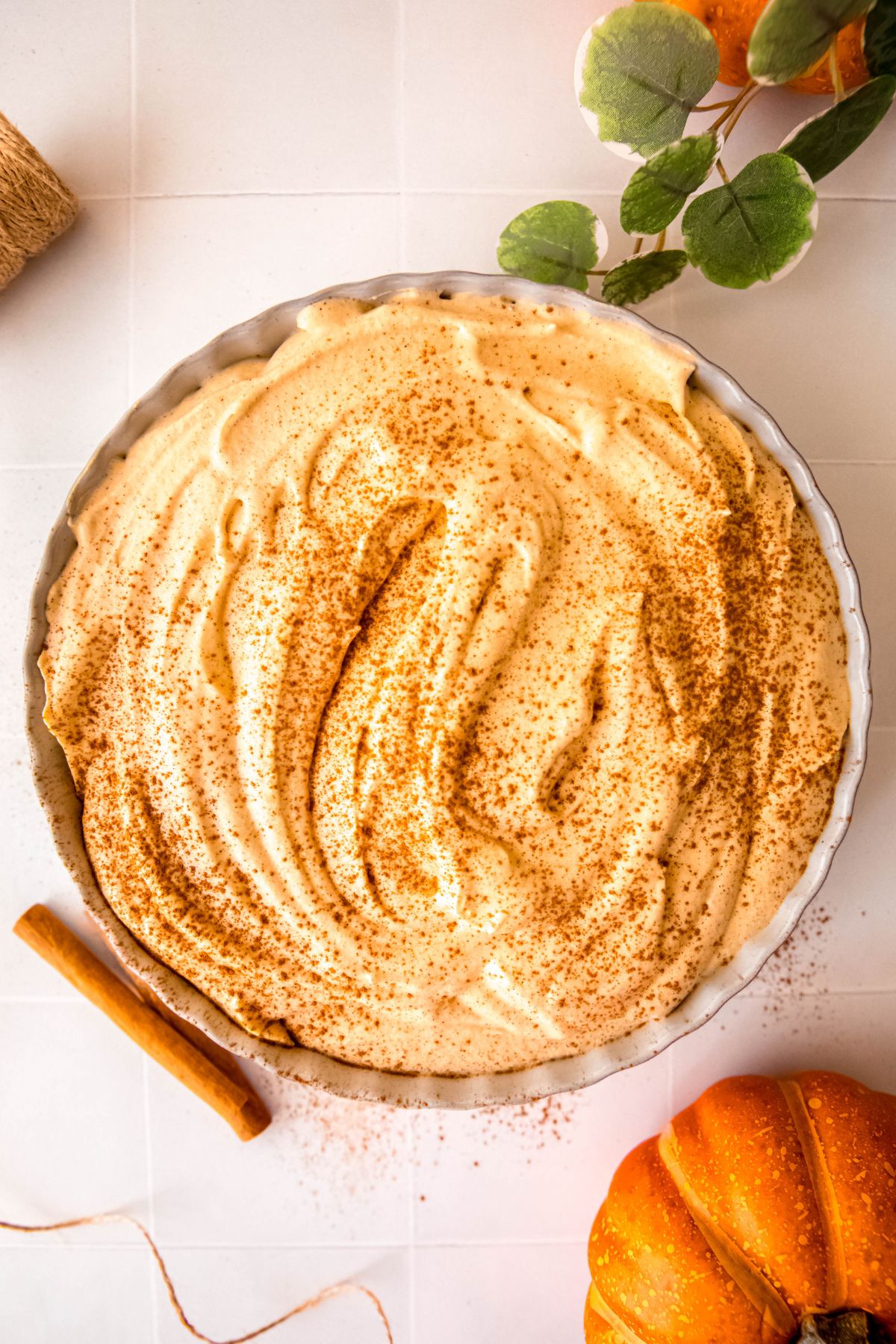 overhead shot of a serving bowl of protein pumpkin fluff dip topped with a sprinkle of cinnamon on a white table with mini pumpkins and cinnamon sticks.