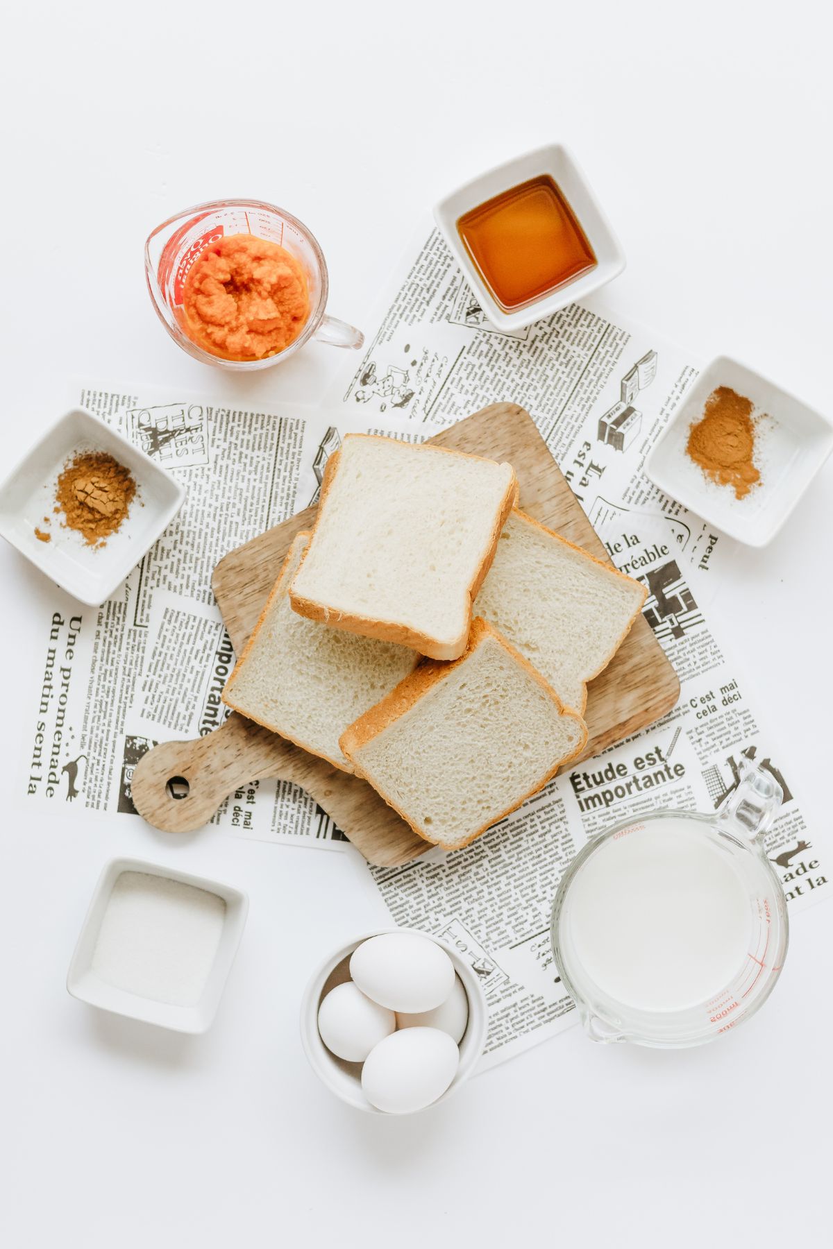 overhead shot of ingredients needed to make pumpkin french toast measured out into white bowls on a white table lined with french newspapers.