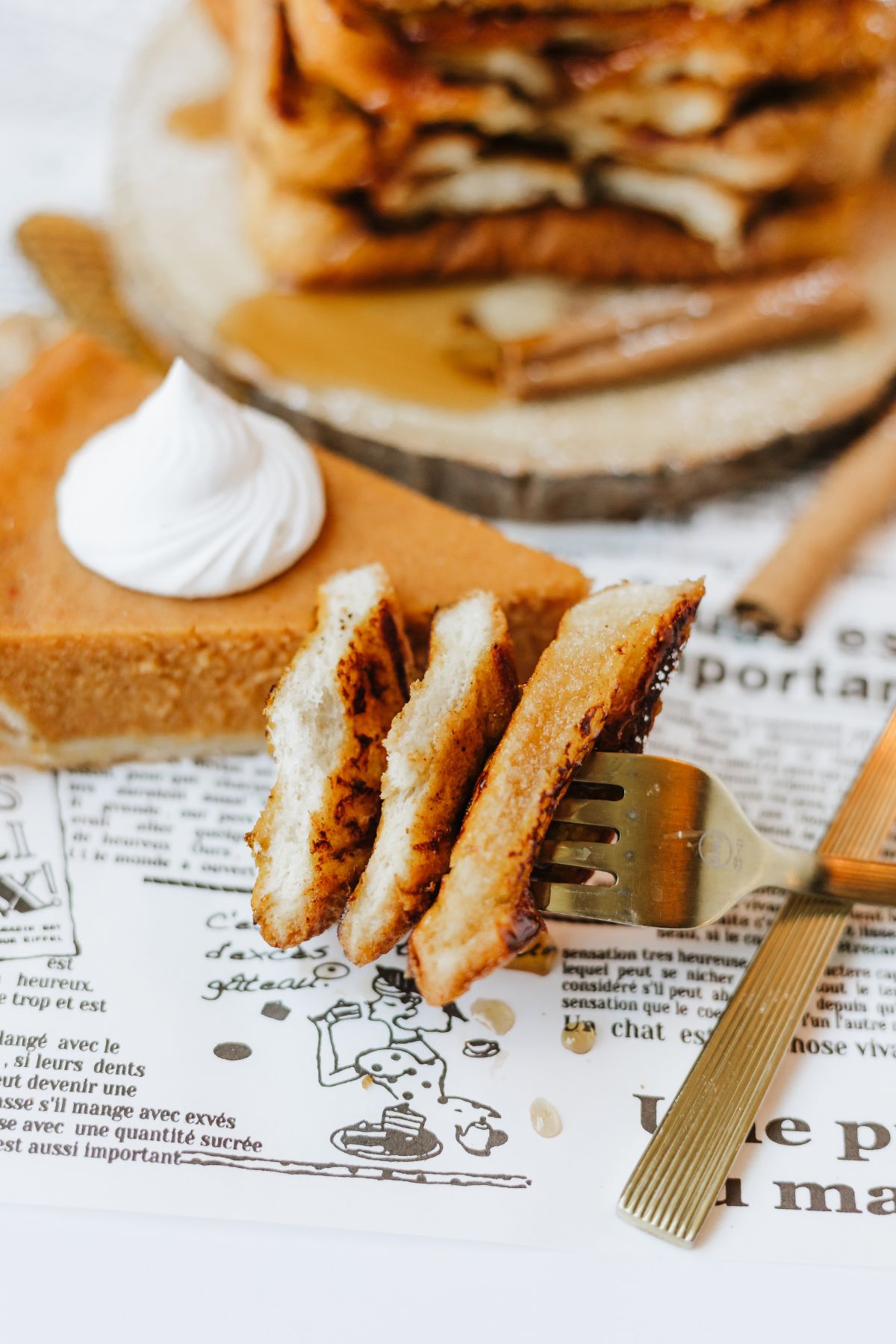 gold fork with a bite cut from 3 pieces of pumpkin french toast dripping a bit of maple syrup onto the french newspaper underneath.