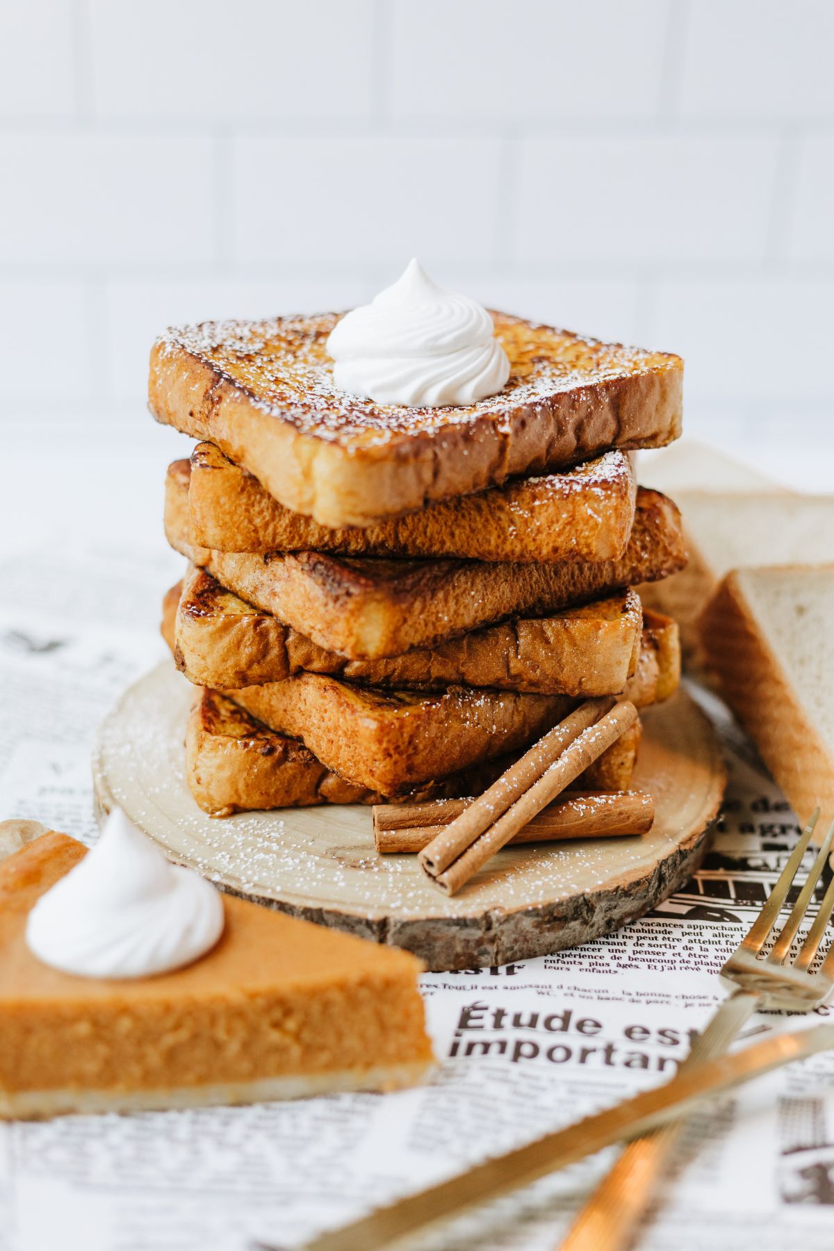 stack of 6 slices of pumpkin spice french toast on a wooden serving plate atop a french newspaper; there's a couple of whole cinnamon sticks leaning against the stack and a slice of pumpkin pie in the foreground.