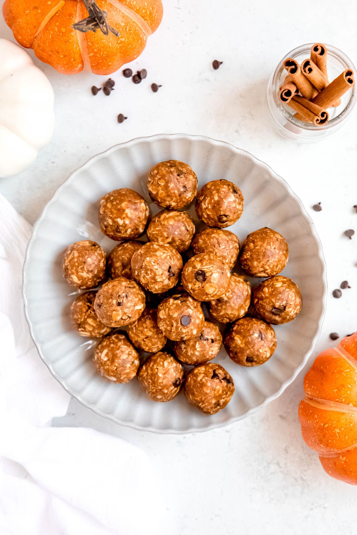 overhead shot of a white, scalloped serving bowl filled with homemade protein pumpkin balls on a white table with mini chocolate chips, two mini pumpkins and a jar with whole cinnamon sticks scattered around.