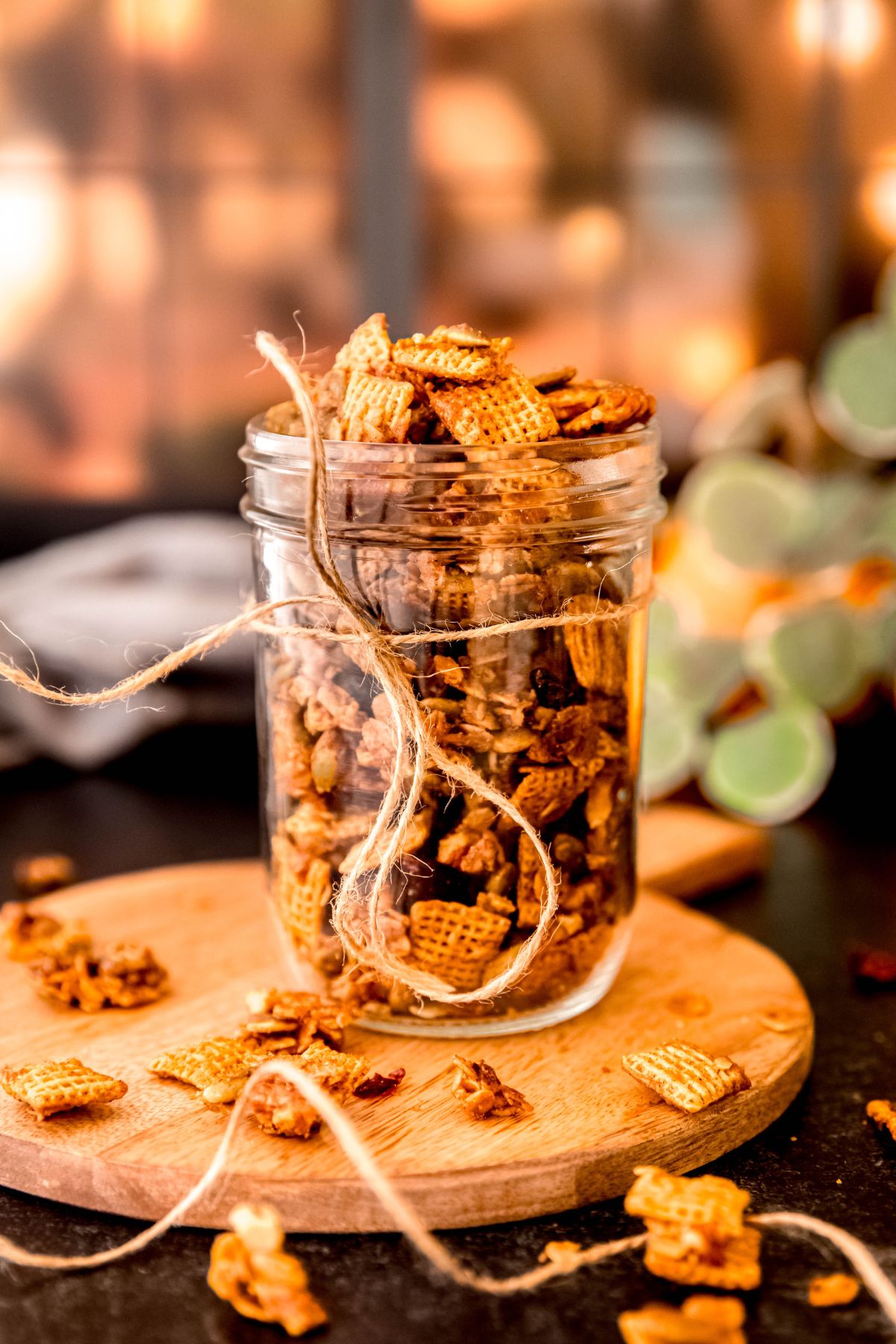 dark countertop with a wooden serving tray topped with a mason jar of pumpkin spice chex mix tied with natural twine in front of a window lit by fairy lights.