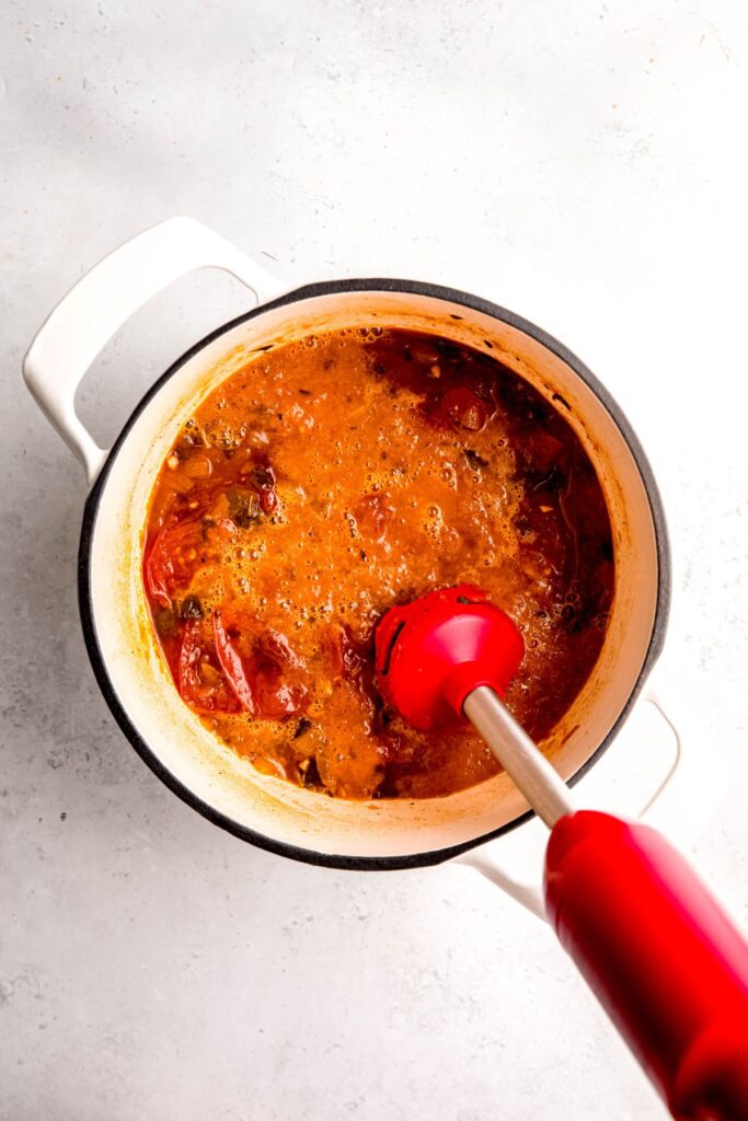action shot of a red immersion blender blending the roasted tomato soup until creamy.