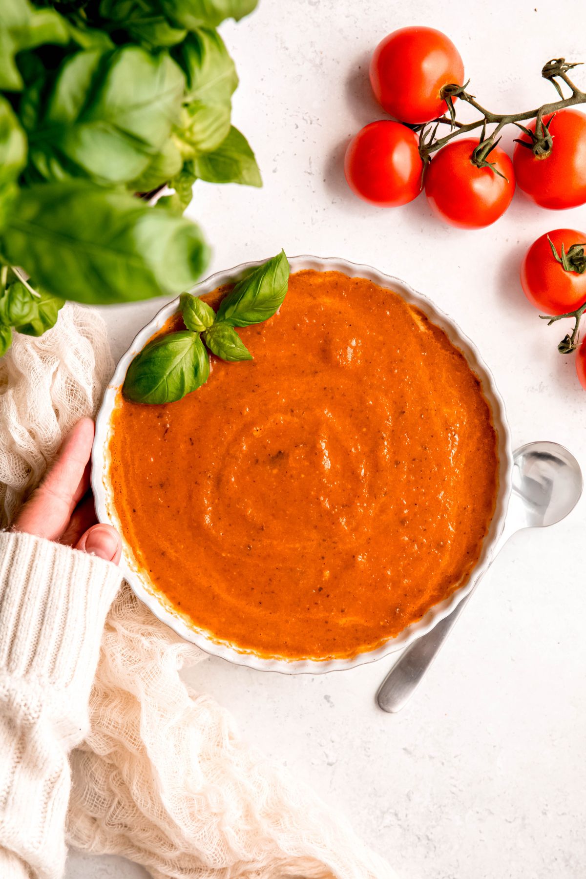 hand in a white sweater setting down a bowl of creamy oven roasted tomato basil soup on a white table with fresh tomatoes and fresh basil.
