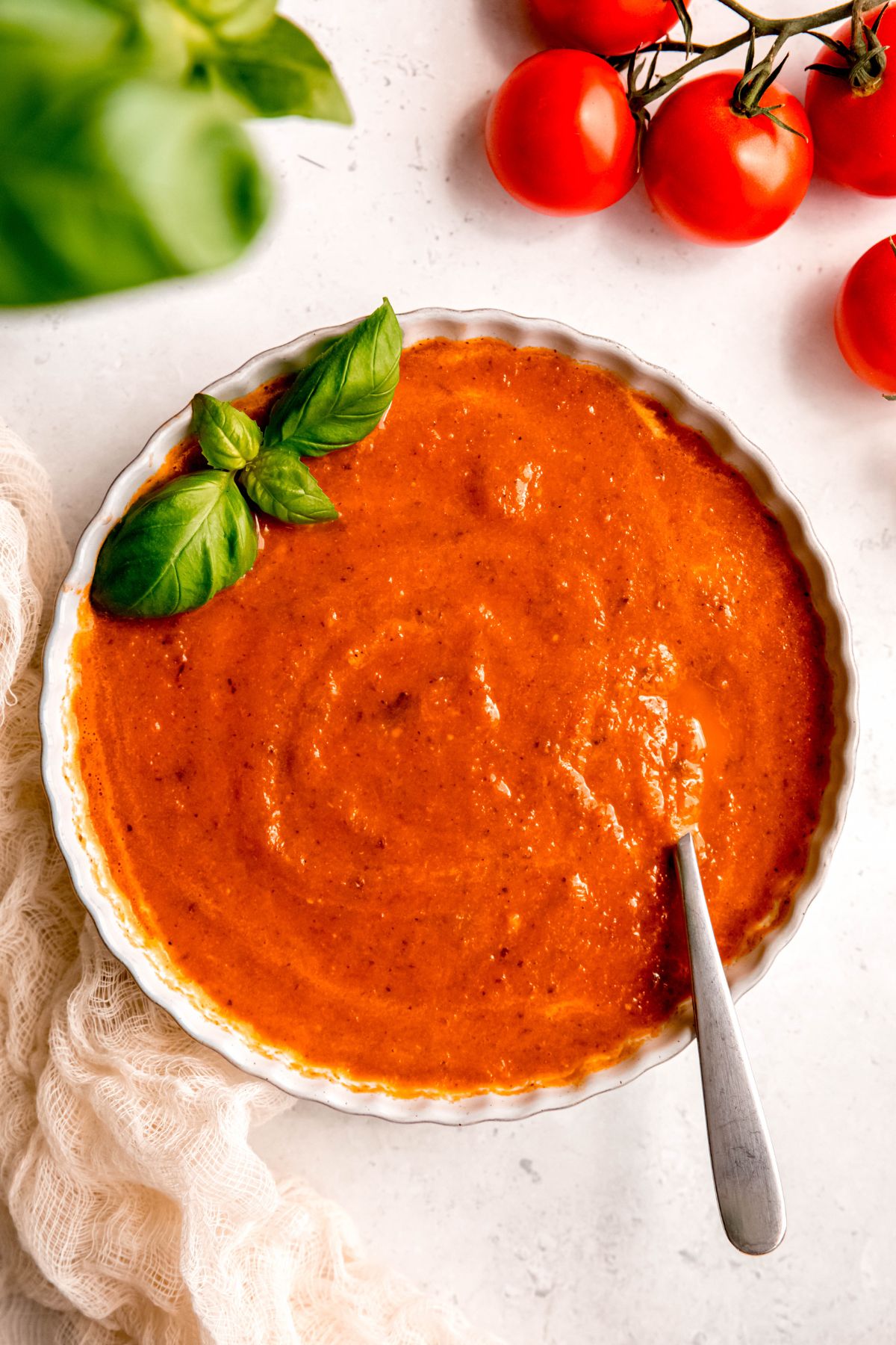 serving of blended roasted tomato soup garnished with fresh basil with a silver spoon sticking out; on the table are more fresh tomatoes and fresh basil.