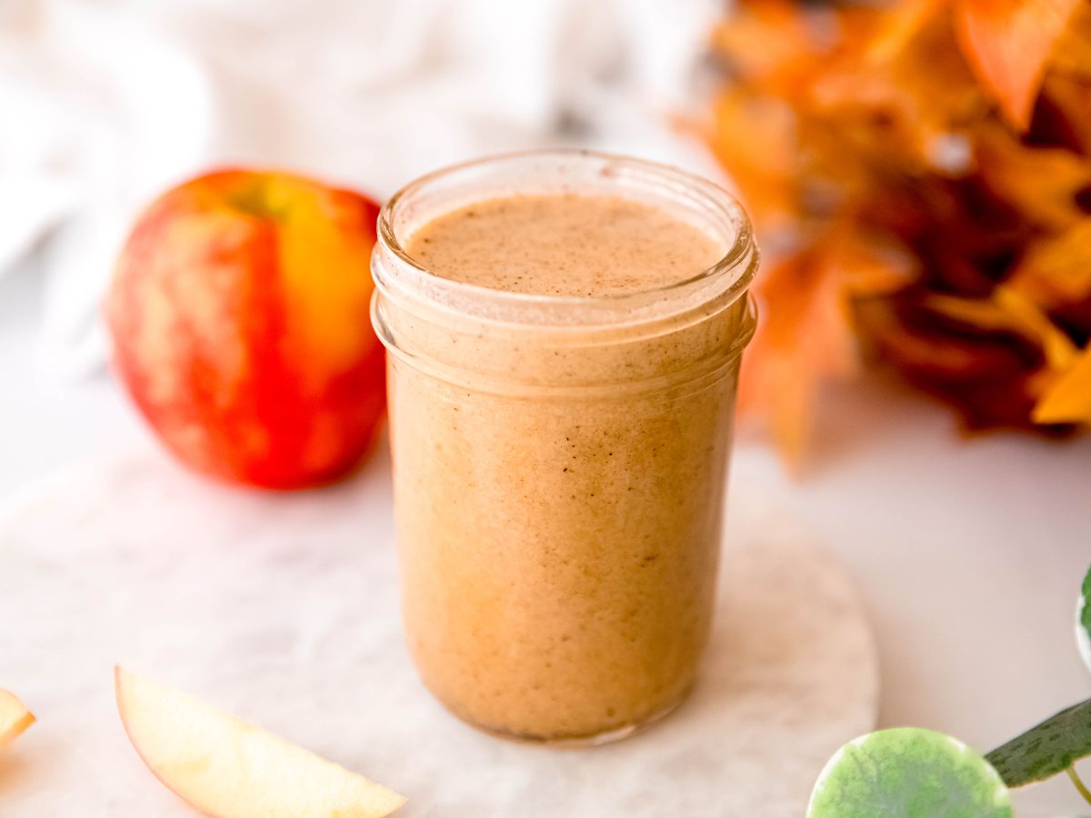 horizontal hero shot of a jar of apple cider salad dressing on a white table with fall leaves and a fresh apple.