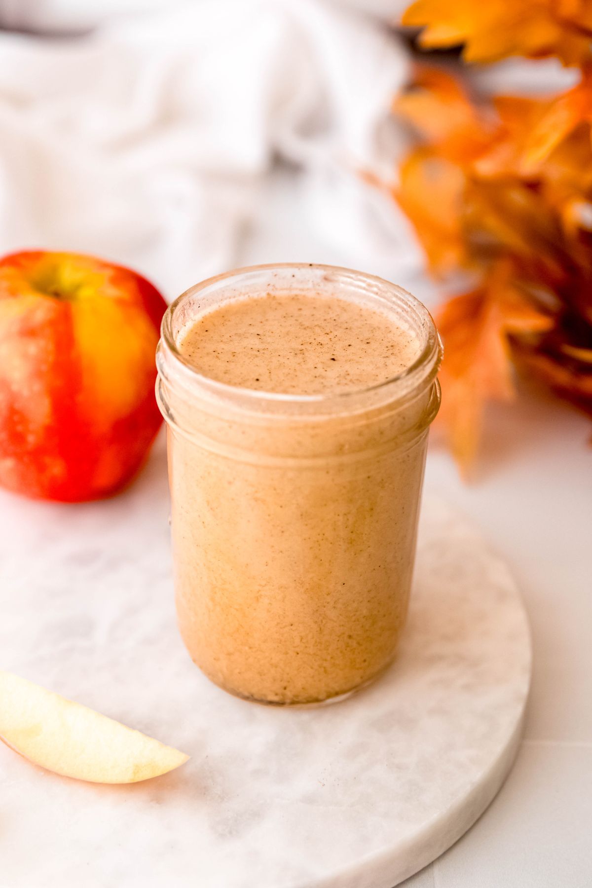 jar of apple cider vinaigrette dressing on a white table with a whole honeycrisp apple, a slice of apple, and a bunch of orange maple leaves.