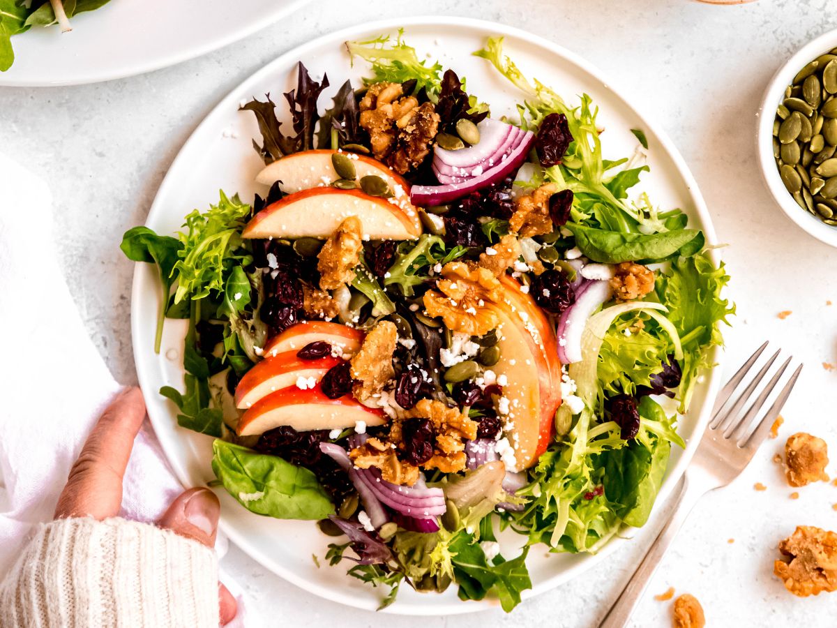 hand setting a salad plate with apple & walnut salad down on a white table with a silver fork.