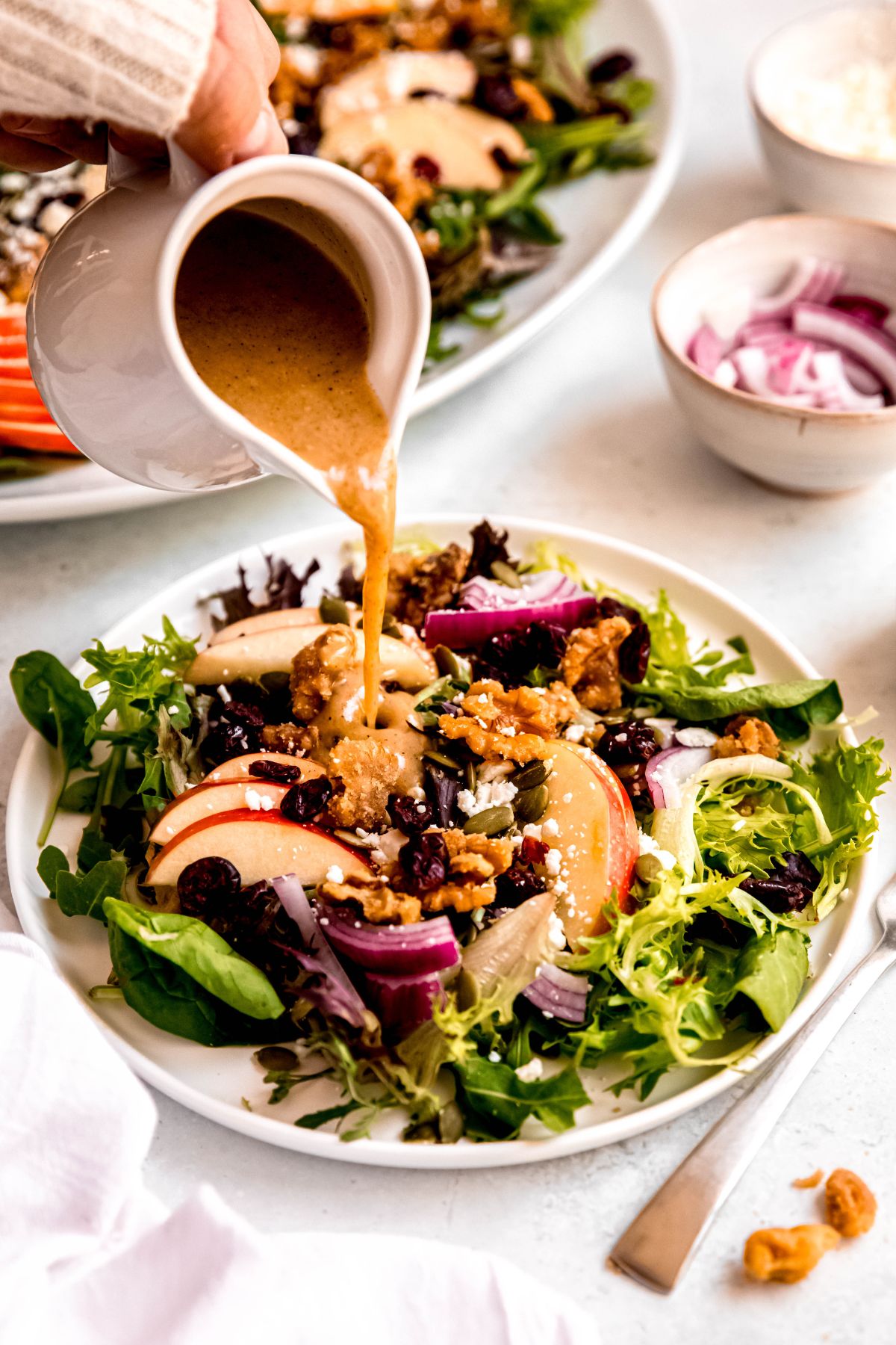 action shot of a hand pouring apple cider vinaigrette from a small white creamer pitcher onto a serving of apple walnut salad with cranberries.