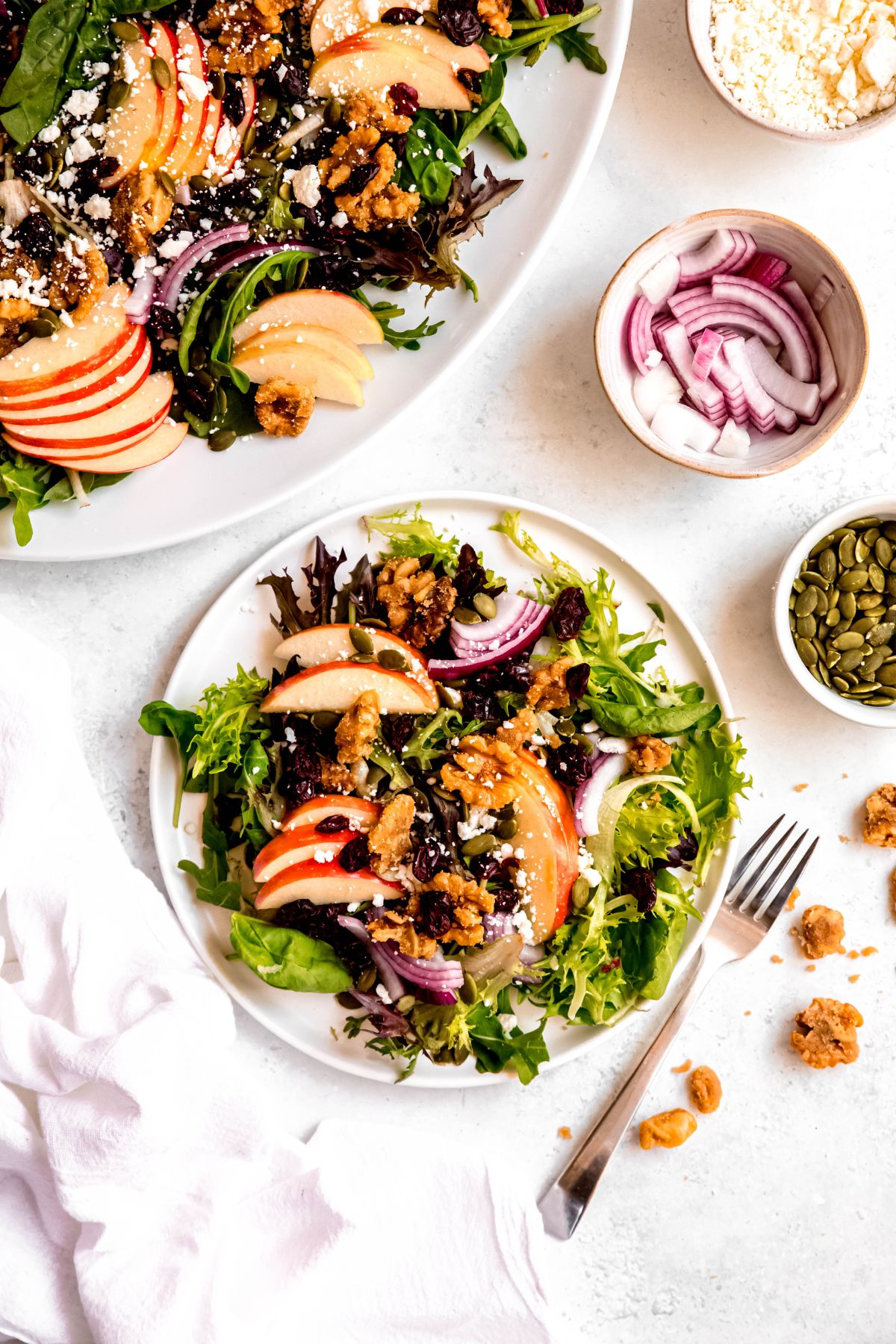 flat lay shot with a white salad plate loaded with a serving of apple and walnut salad on a white table with the serving platter with the rest of the salad and small bowls with extra sliced red onions, pepitas, and candied nuts framing the shot.