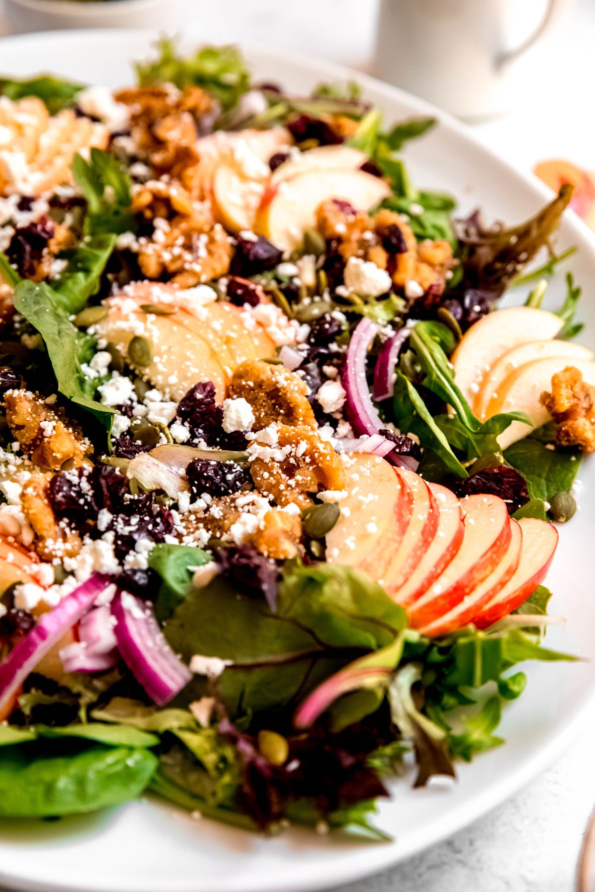 closeup shot of an apple and walnut salad showing the greens on the bottom topped with apples, candied nuts, pepitas, dried cranberries, red onions, and feta crumbles.