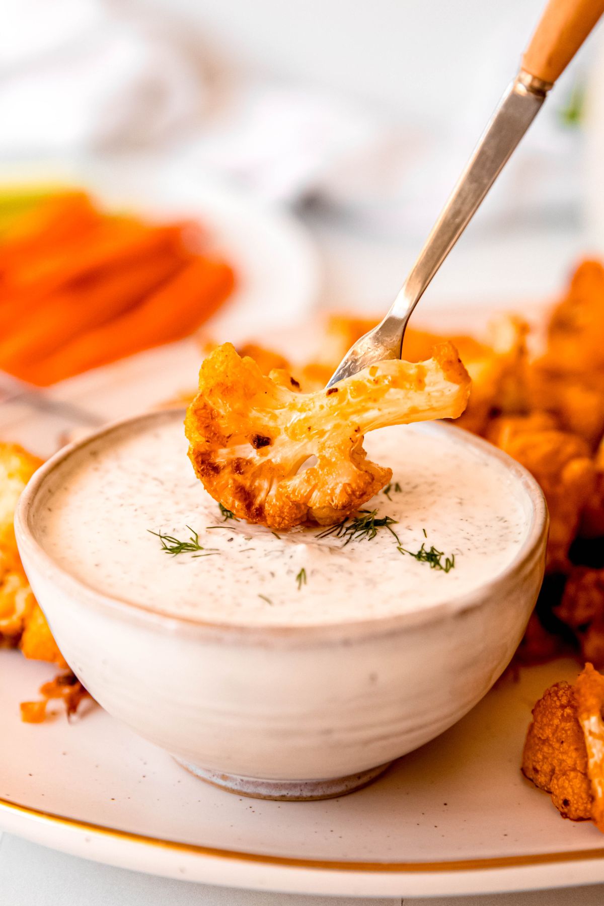 closeup side on shot of a spicy, crispy buffalo cauliflower wing bite being dipped into the ranch dressing bowl using an oyster fork.