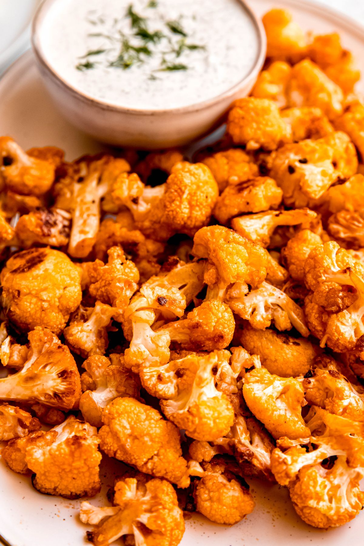 closeup shot of a pile of roasted baked cauliflower buffalo wings next to a bowl of ranch dressing garnished with fresh herbs.