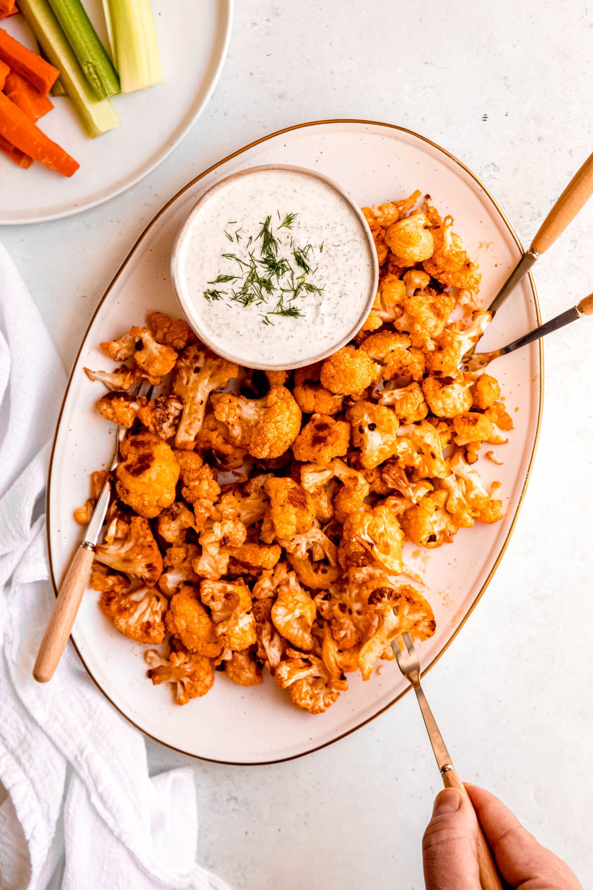 overhead shot of a serving platter of oven baked buffalo cauliflower wing bites with silver and wooden oyster forks for serving and a white bowl filled with greek yogurt ranch dressing sprinkled with fresh dill.