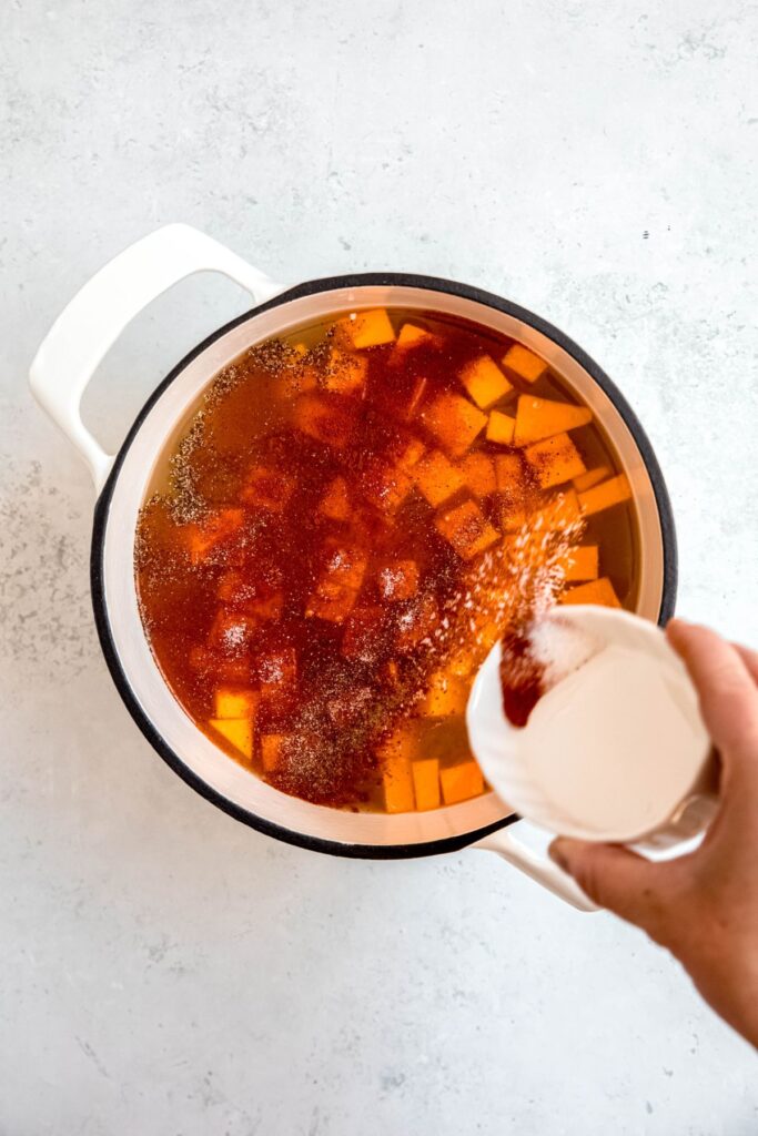 action shot of a hand sprinkling the seasoning mix over the pot after adding the remaining broth and butternut squash cubes.