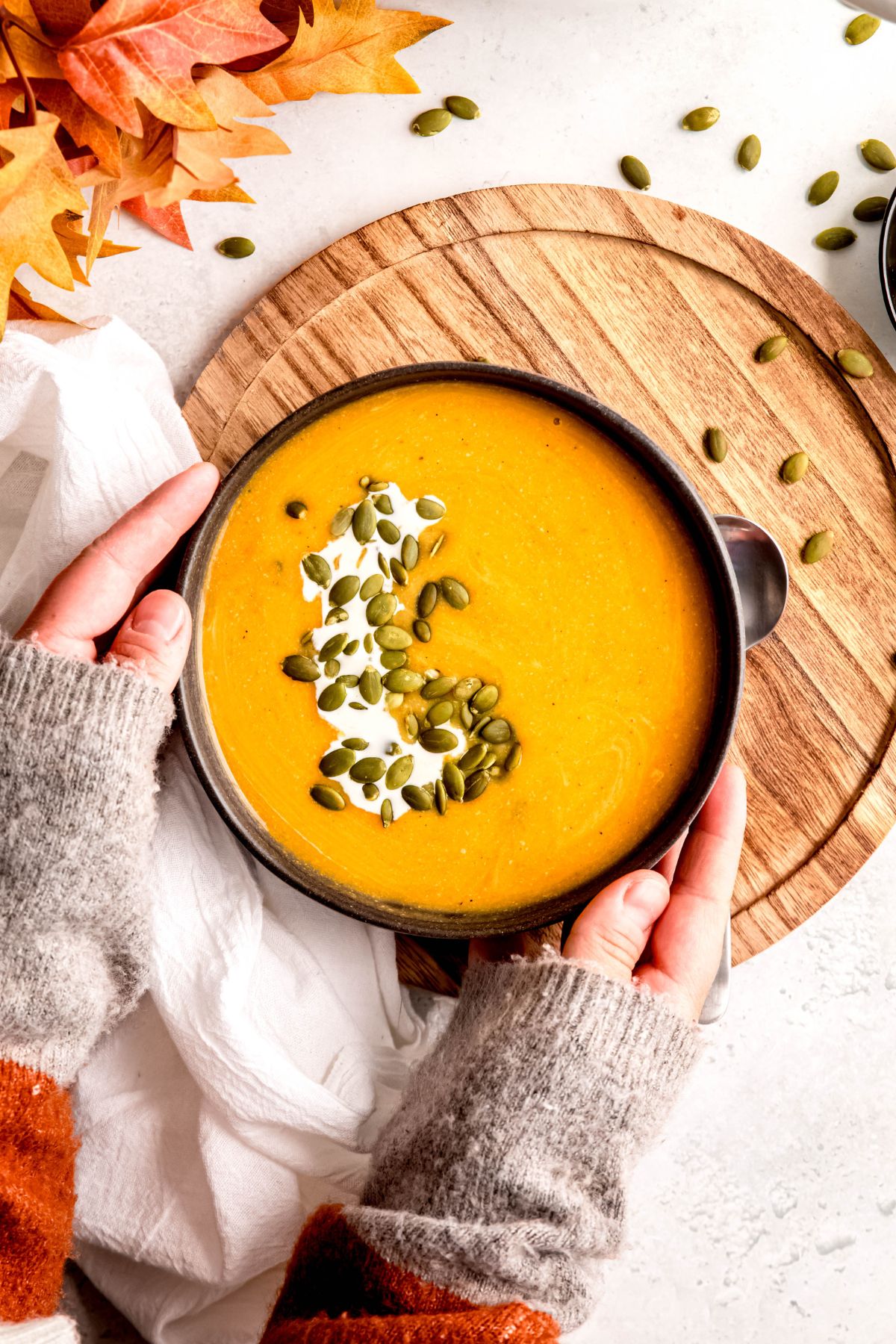 hands in a grey sweater setting down a garnished bowl of creamy high protein butternut squash soup on a wooden charger on a white table with red/orange fall leaves and pepitas scattered across the table.