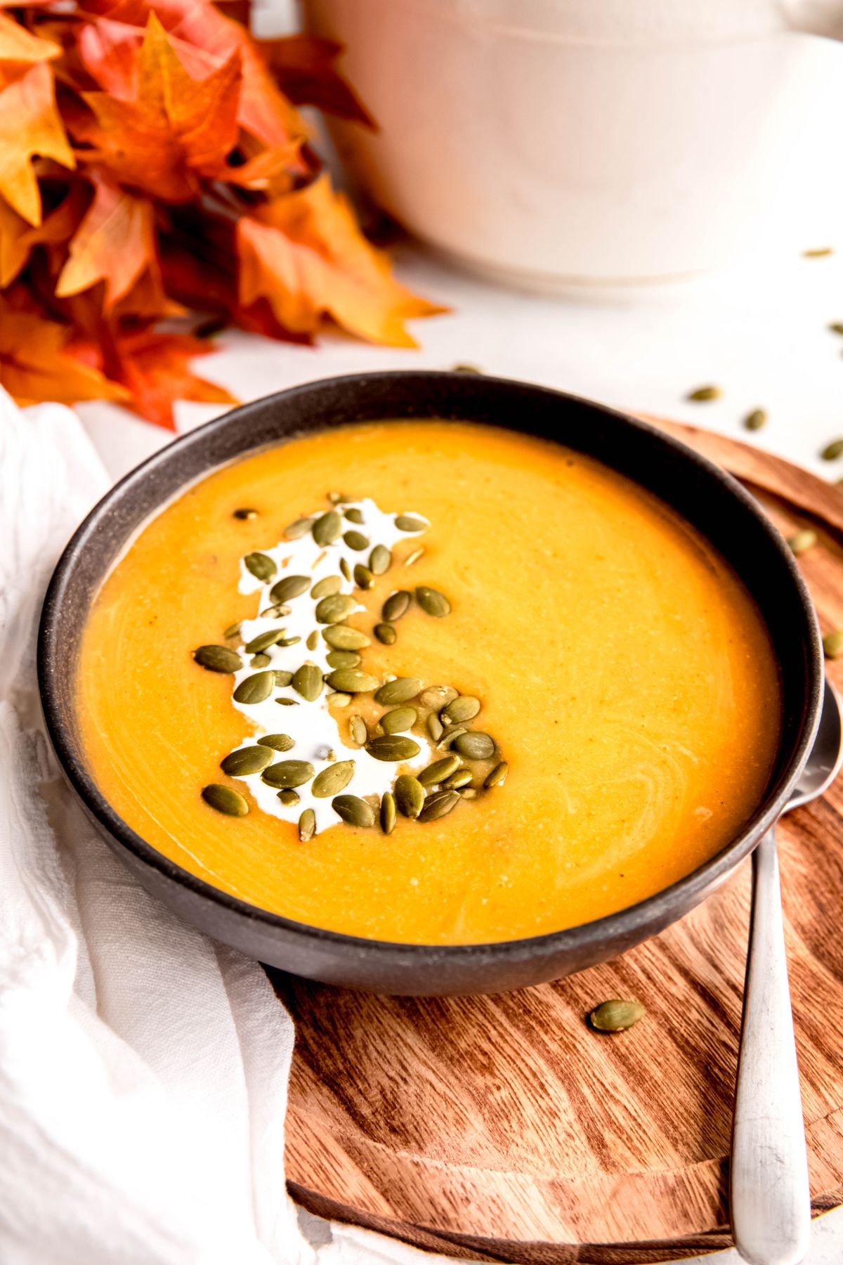 cozy fall table decorated with fall leaves, white linen, and a wooden charger with a bowl of protein-packed creamy apple butternut squash soup garnished with a drizzle of cream and sprinkle of roasted pepitas.