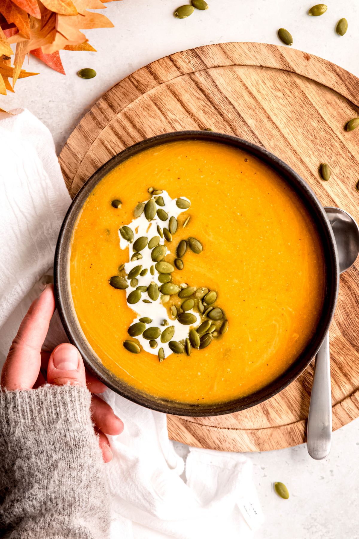 left hand clad in a cozy grey sweater is resting against the left side of the butternut squash and apple soup bowl as if it had just been set down.