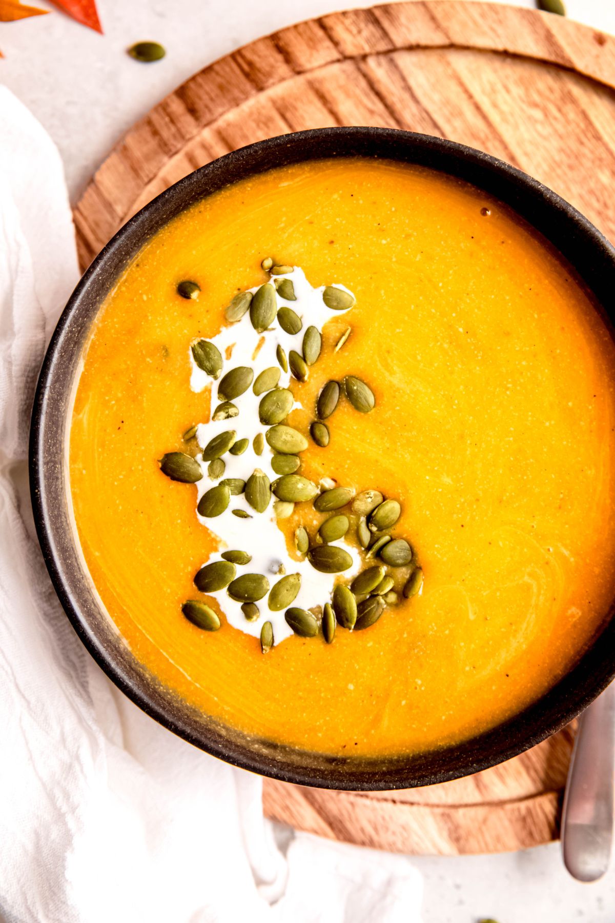 closeup shot of a bowl of creamy butternut squash protein soup garnished with pepitas and a drizzle of cream on a wooden cutting board with a clean white kitchen towel to the side.