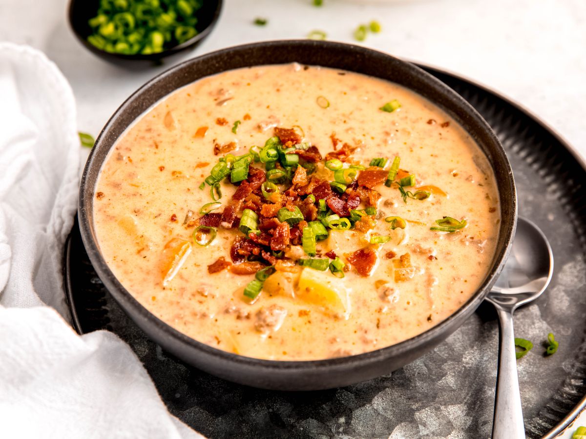 horizontal hero shot of a bowl of creamy cheeseburger soup on a black charger with a silver spoon on a white table with a small black pinch bowl of dark green sliced scallions.