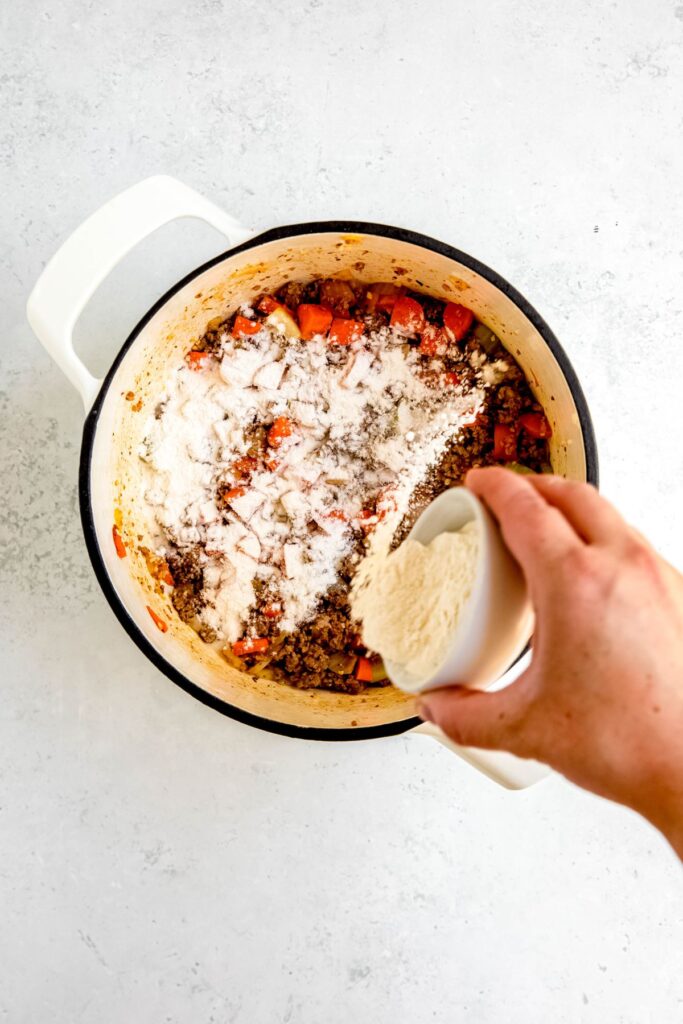 action shot of a hand sprinkling the flour into the pot to cook with the meat and veggies.