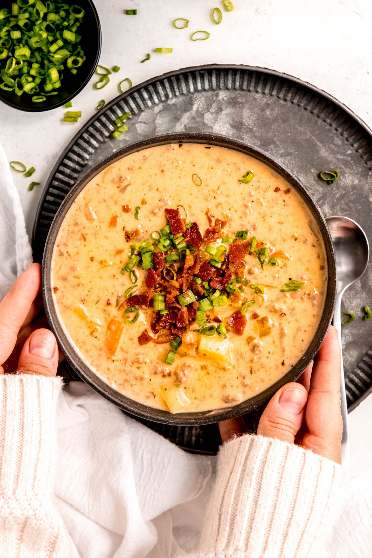 hands in a cozy white sweater placing a bowl of homemade creamy cheeseburger soup on a black metal charger on a white table.