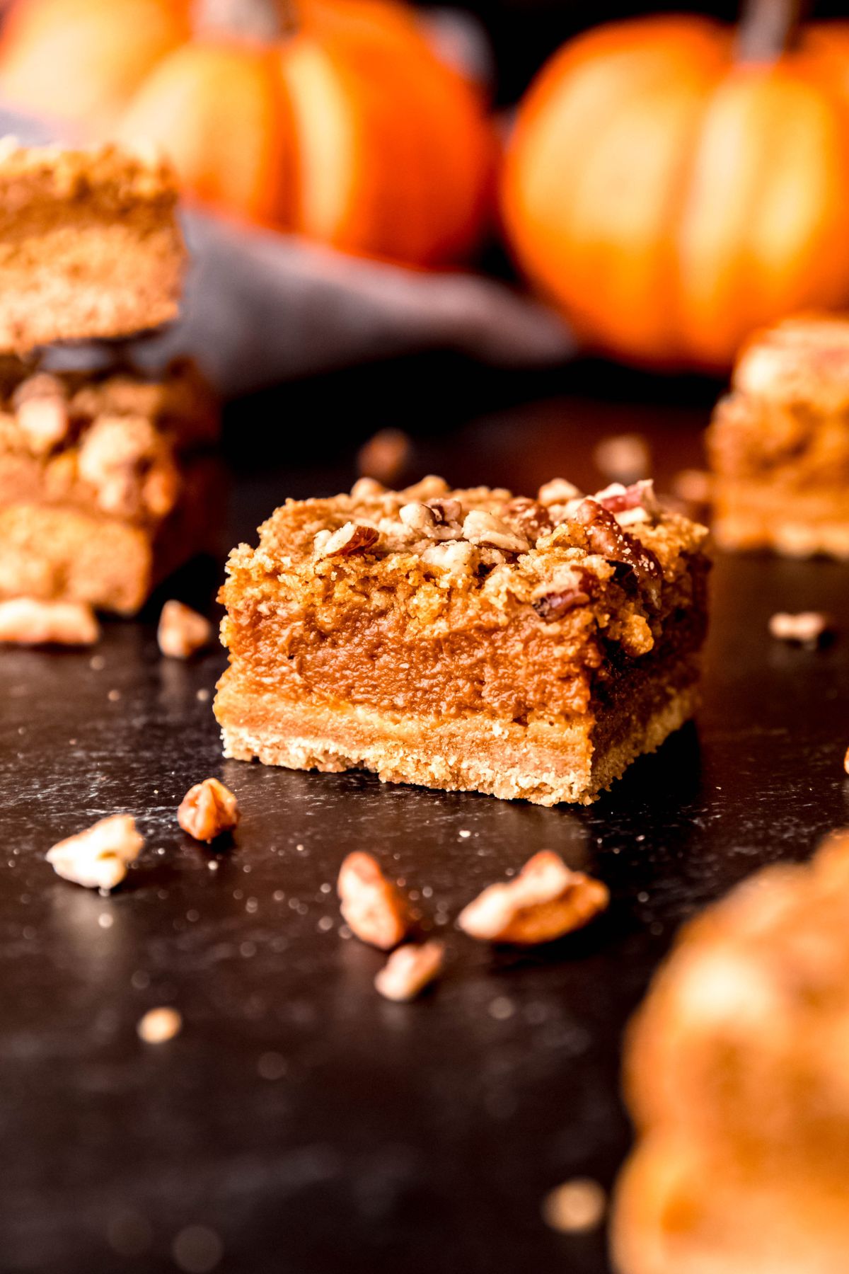 side on shot of a table with pumpkin crumble bars and chopped pecans scattered around with just one crumb bar in focus in the middle.