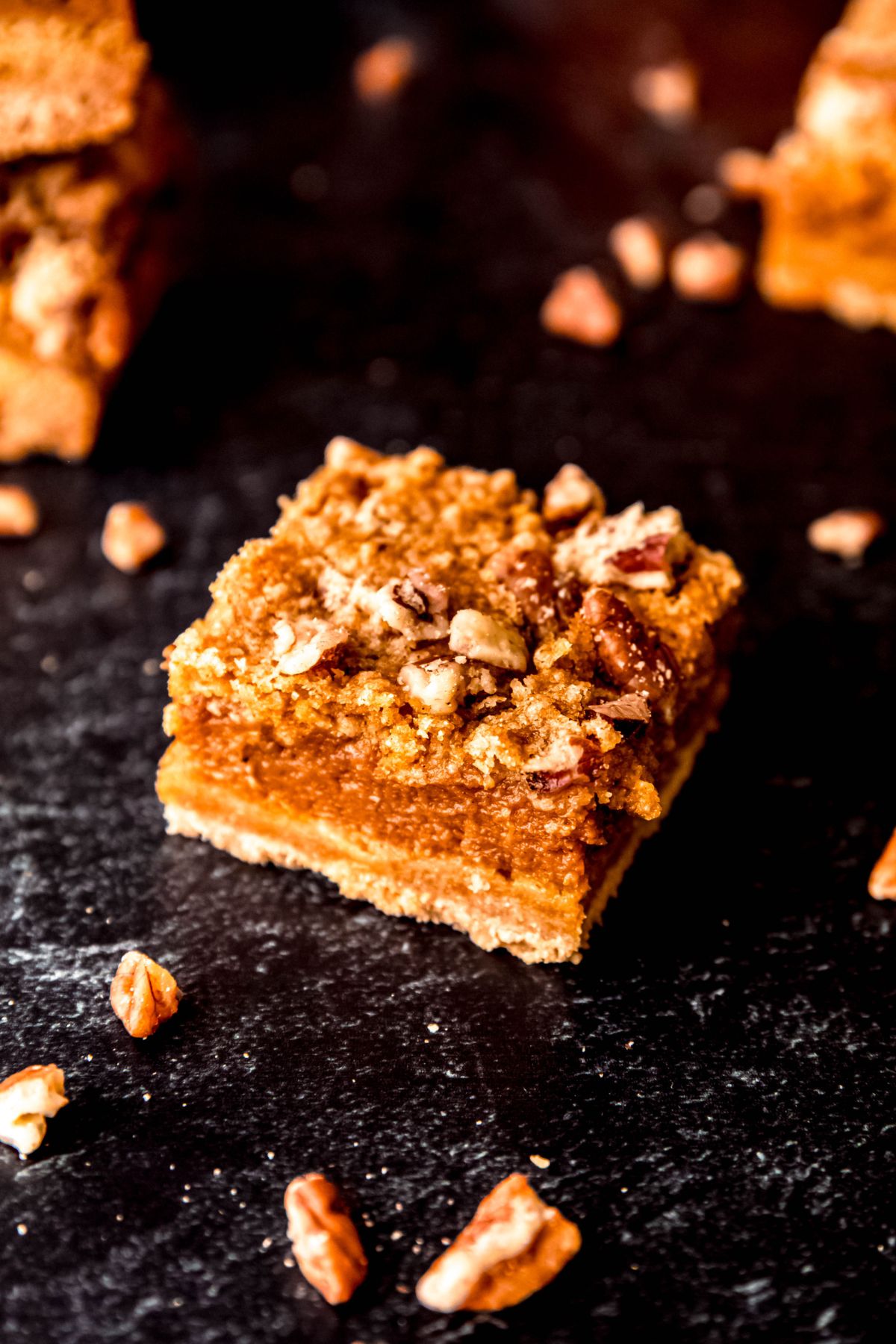 closeup of a single pumpkin crumb bar on a black table with chopped pecans scattered around.