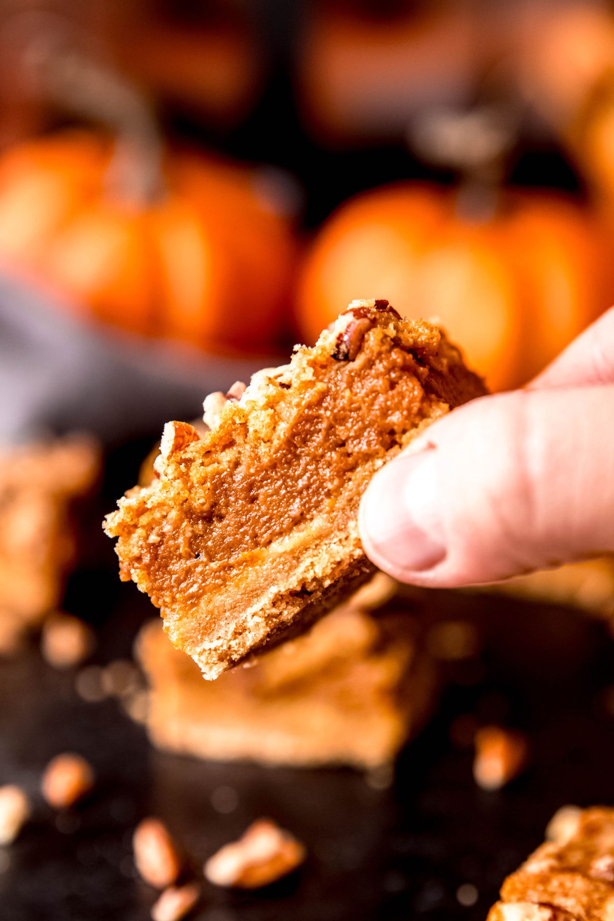 hand holding the corner of a pumpkin crumble bar above a black table with the remaining bars and pecans scattered around blurred out below.