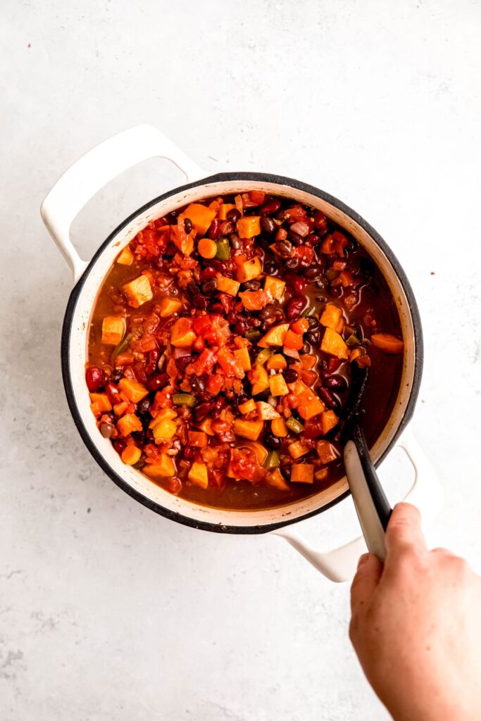 A hand stirring a pot of veggies, beans, seasoning, and canned tomatoes.
