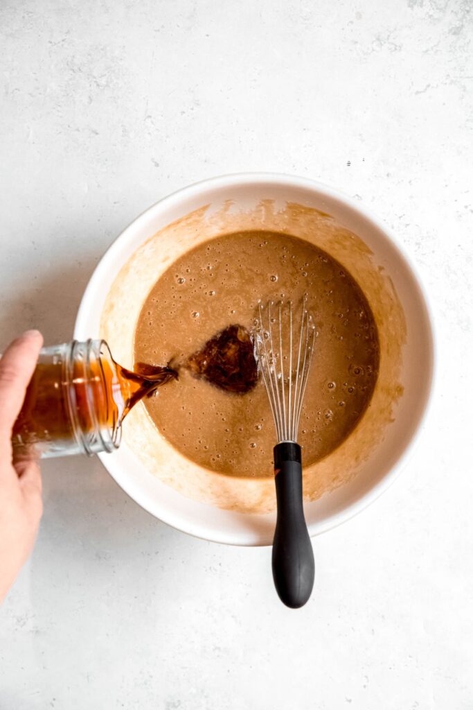 action shot of a hand pouring the cooled reduced apple cider into the bowl with the wet ingredients and sugar.