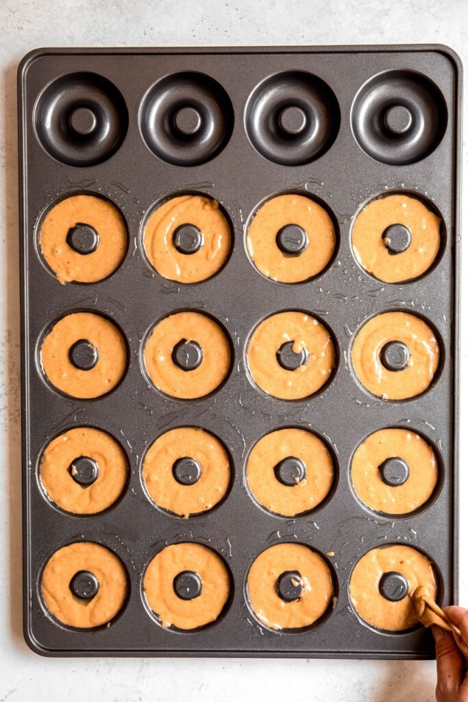 action shot of a hand piping the apple donut batter into a doughnut pan.