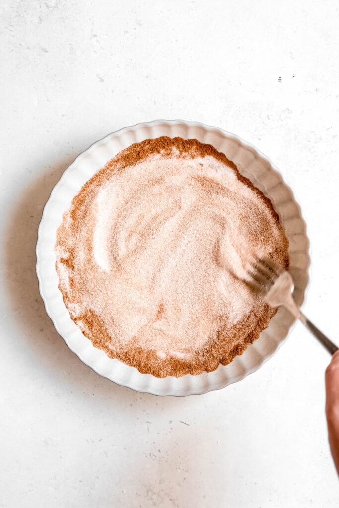 action shot of a fork stirring together cinnamon and sugar in a wide, shallow bowl.
