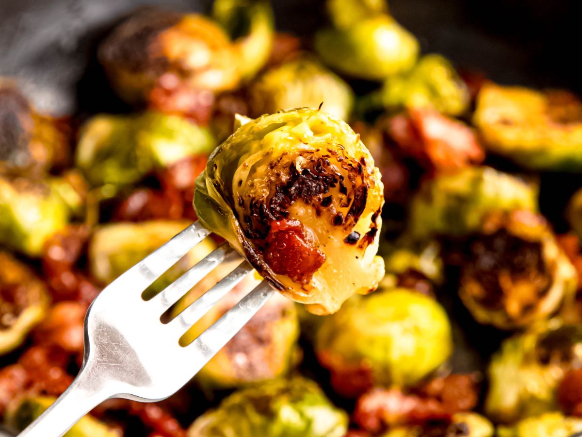 horizontal rectangular hero shot of a silver fork with a dark golden brown bite of brussels sprouts with bacon bits in front of a blurred out pan filled with the rest of the batch.