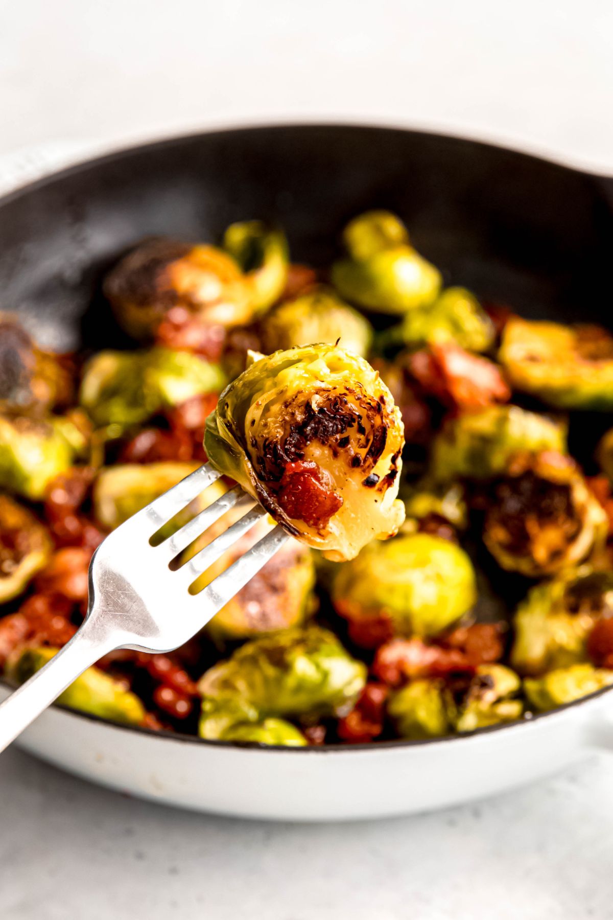 silver fork with a caramelized brussels sprout with bacon bits clinging to it with the rest of the batch of sprouts in a skillet blurred out in the background.
