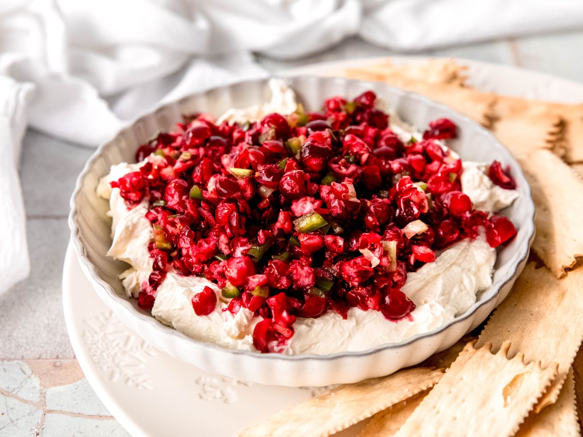 horizontal rectangular hero of cranberry and jalapeno dip with whipped cream cheese in a bowl on top of a plate with crackers.